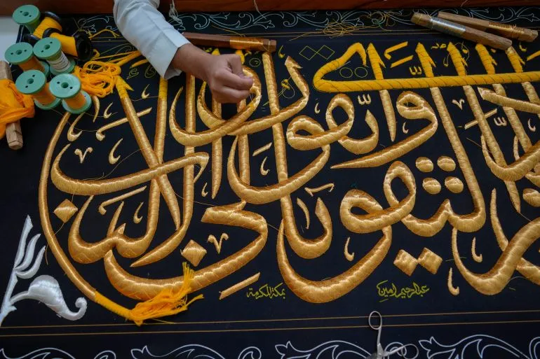 تفاصيل دقيقة عن التطريز الذهبي على الكسوة السوداء للكعبة، مع وجود خيوط ملونة وأدوات الخياطة، تعكس الفنون الإسلامية التقليدية.