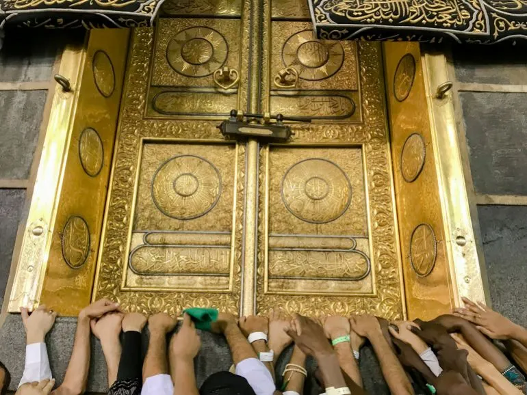 أيدي الحجاج تتجمع حول الباب الذهبي للكعبة المشرّفة، مع وجود الكسوة السوداء المزخرفة في الأعلى، تعبيراً عن الإيمان والعبادة.