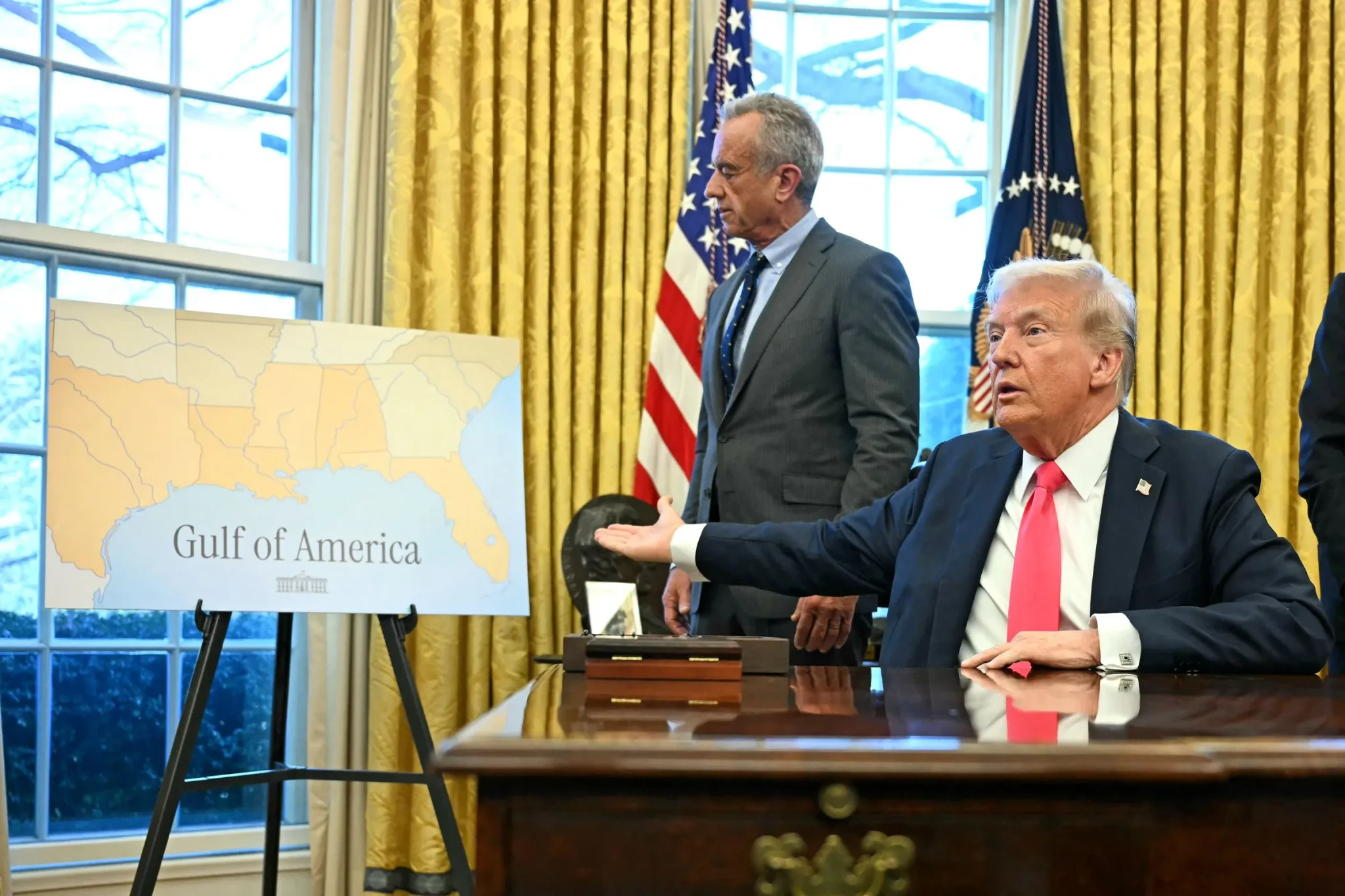 الرئيس الأمريكي دونالد ترامب يجلس على مكتب في المكتب البيضاوي ويشير إلى خريطة تُظهر إعادة تسمية خليج المكسيك إلى "خليج أمريكا" ضمن حملة تغيير أسماء جغرافية لتعزيز الهوية الأمريكية.