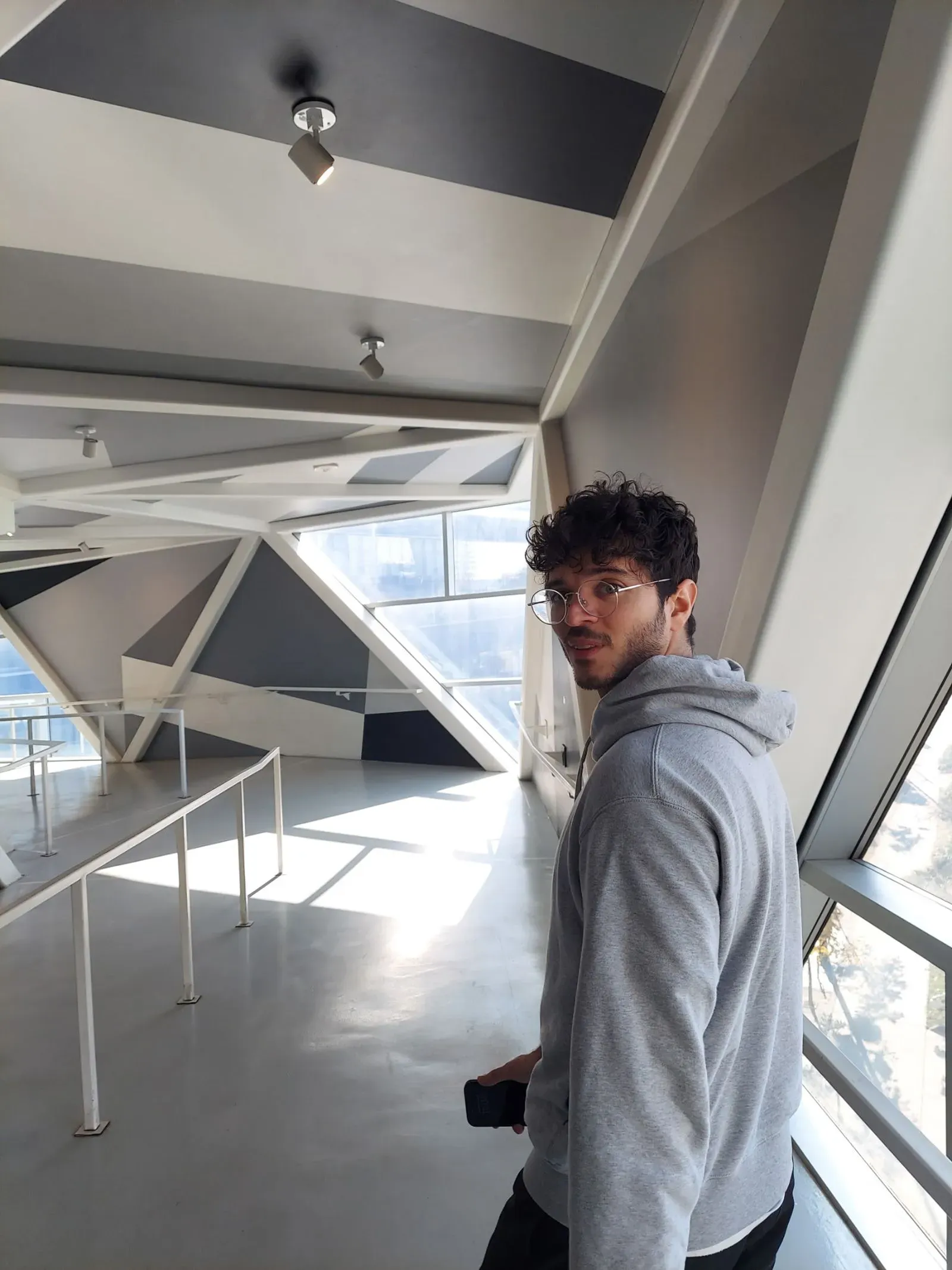 شخص يرتدي سترة رمادية يتجول في ممر داخلي حديث بتصميم معماري متميز، مع نوافذ كبيرة تسمح بدخول الضوء الطبيعي.