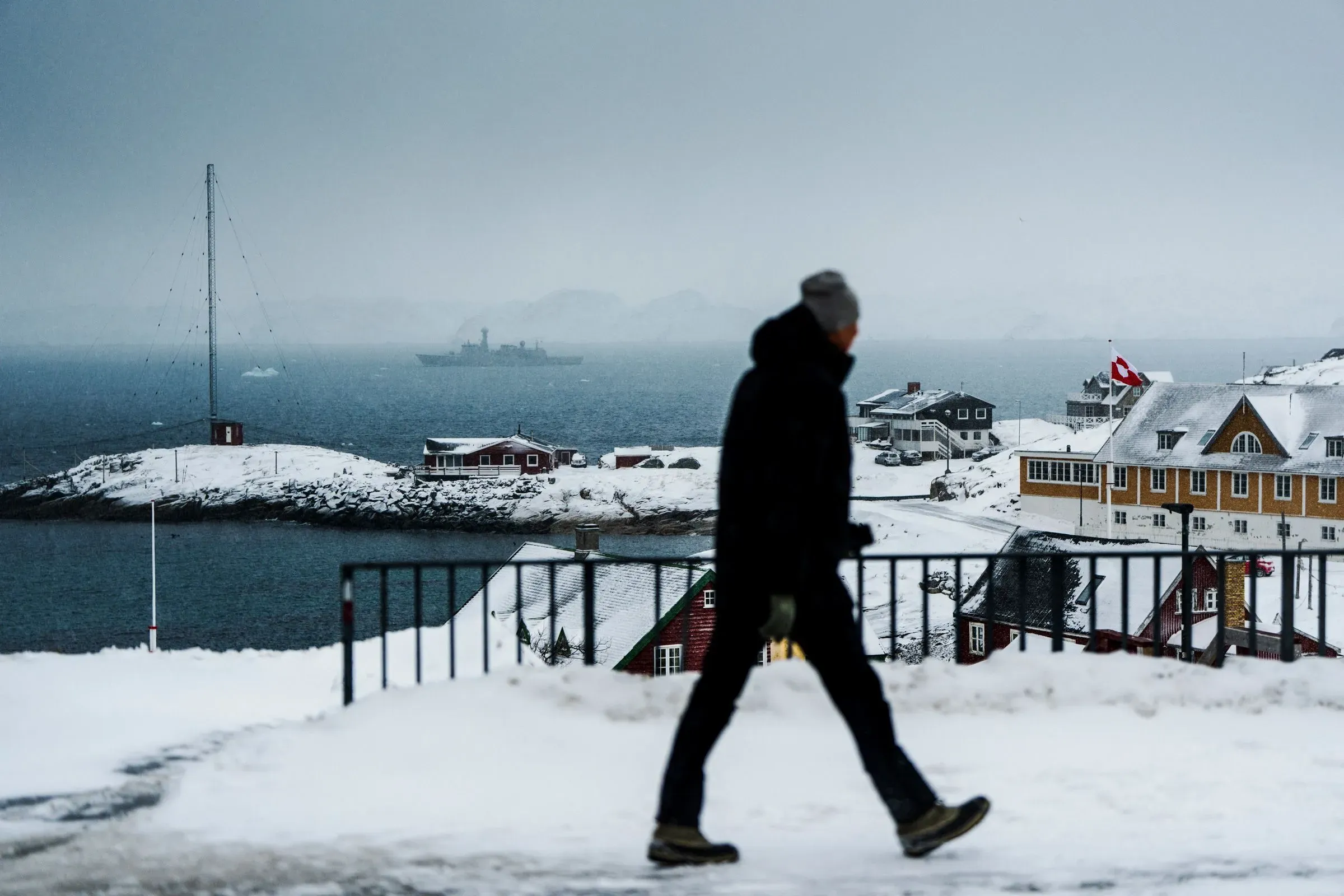 مشهد لمدينة غرينلاند المغطاة بالثلوج، حيث يسير شخص في ملابس شتوية، مع سفينة في البحر وخلفية من المنازل الملونة.