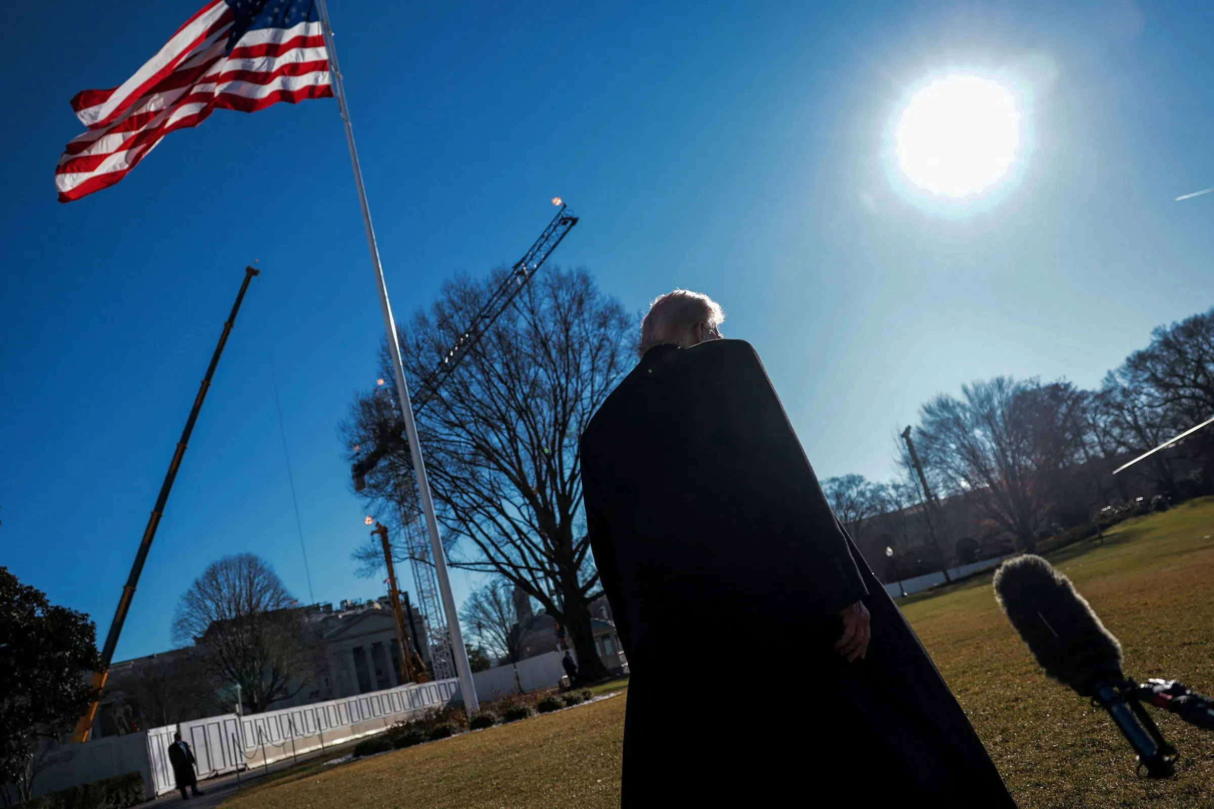 صورة لرجل يقف في حديقة البيت الأبيض، مع العلم الأمريكي يرفرف في الخلفية، تحت سماء زرقاء مشمسة. تعكس الصورة التوترات السياسية الحالية.