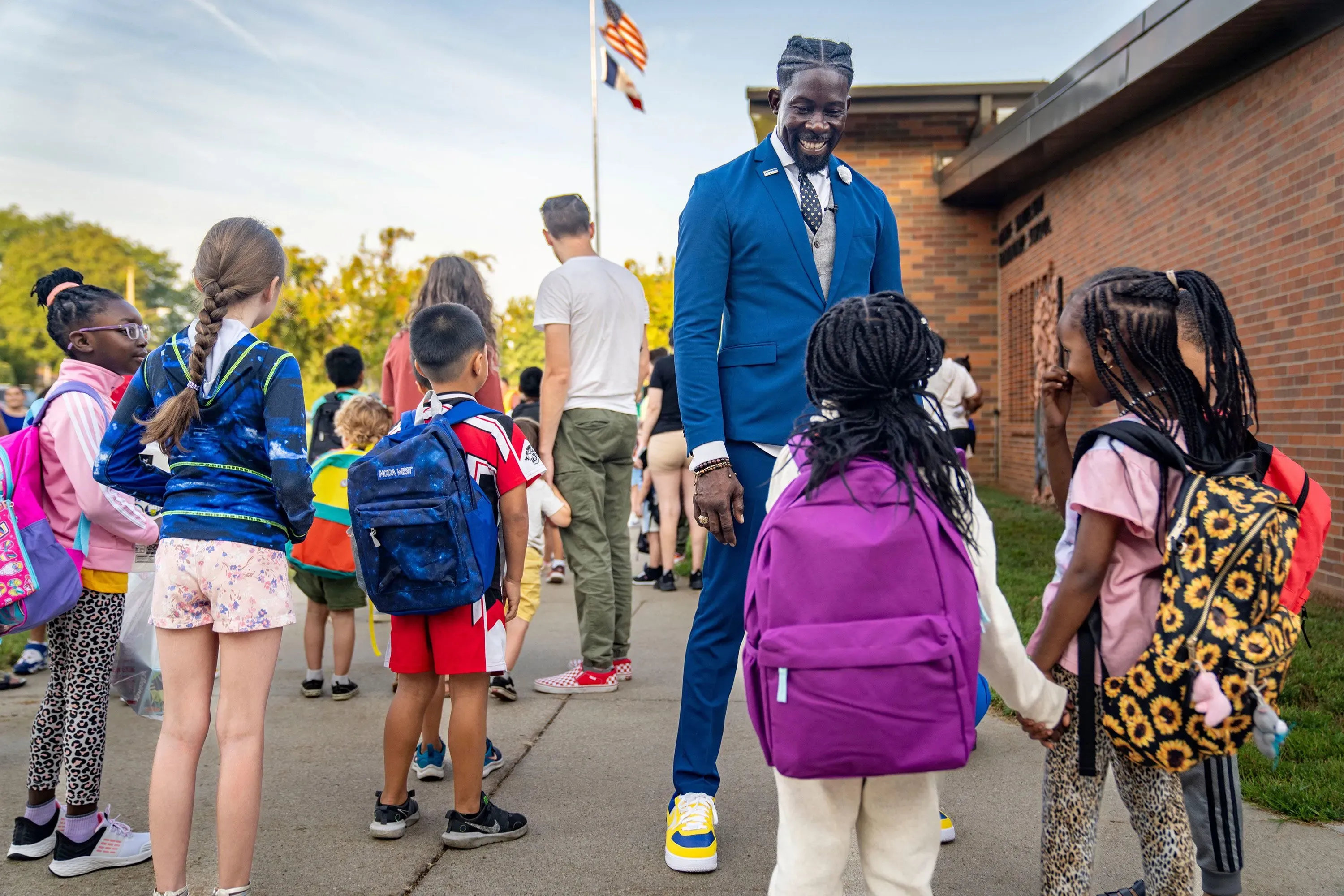مدير مدارس دي موين، إيان روبرتس، يتفاعل مع مجموعة من الأطفال في ساحة المدرسة، مرتديًا بدلة كستنائية وحذاء رياضي، مما يعكس روح التعليم والمشاركة.