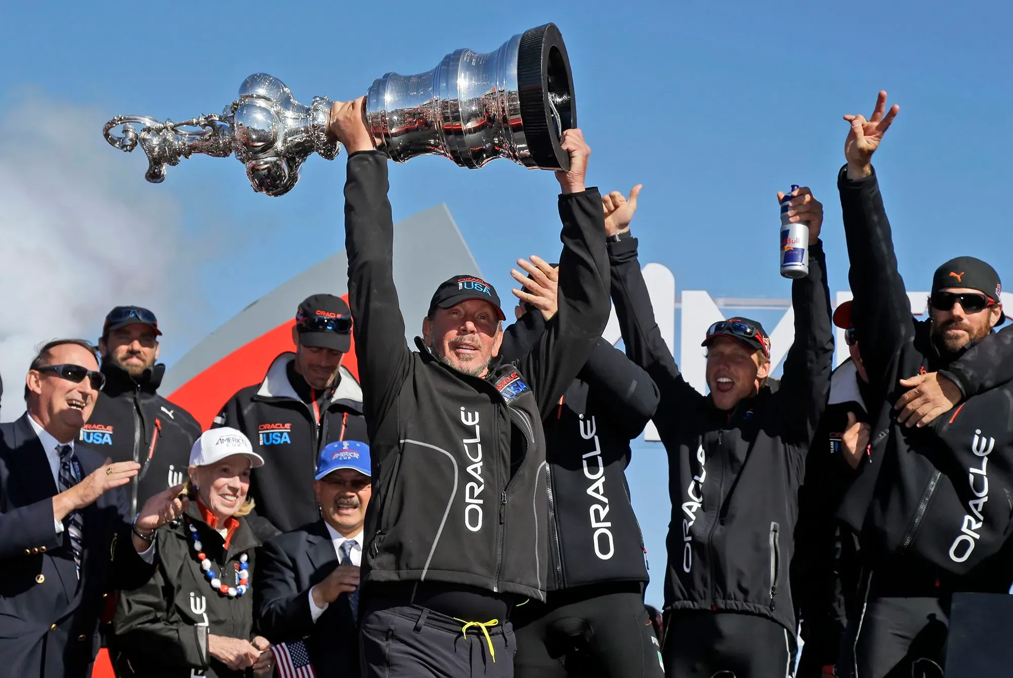 فريق أوراكل يرفع كأس أمريكا في احتفال انتصاره، مع وجود لاري إليسون في المقدمة، محاطًا بفرقة من أعضاء الفريق.