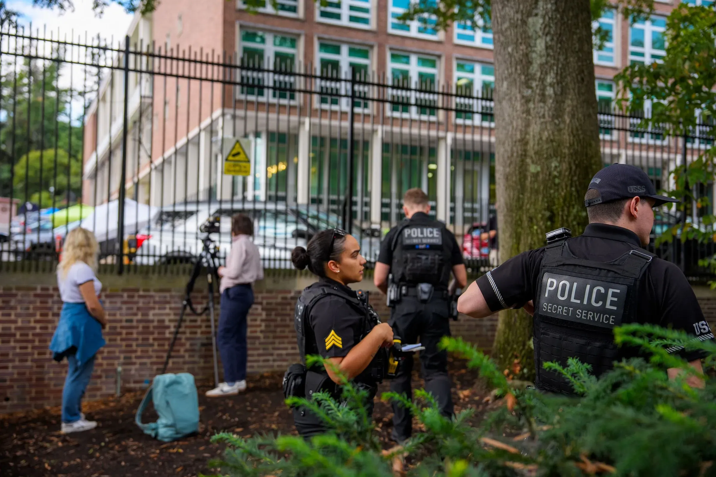 وجود عناصر من الشرطة السرية أمام مبنى، مع تصوير لعدد من الأشخاص في الخلفية، مما يعكس الأجواء المحيطة بالتحقيقات الجارية.