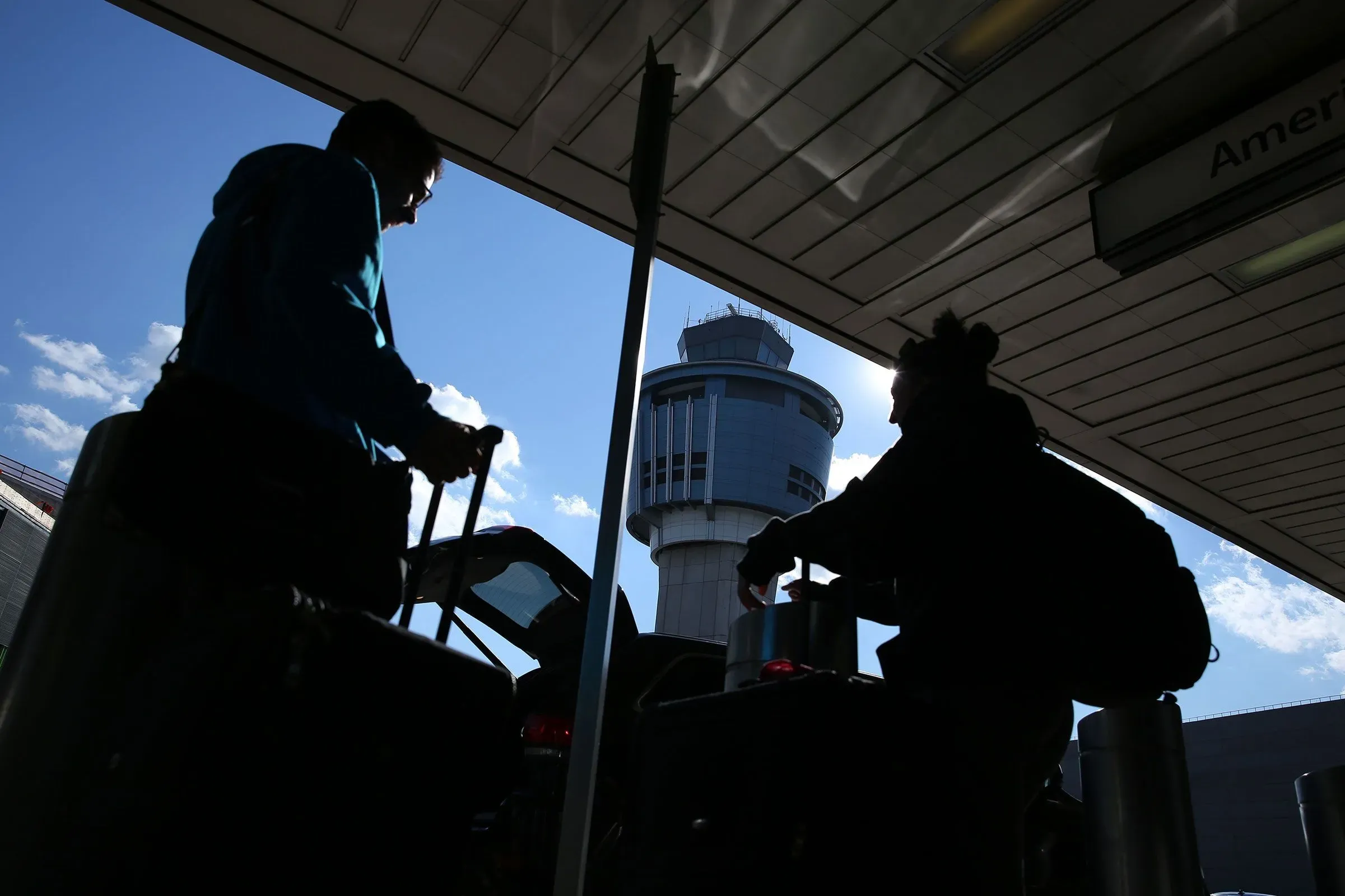 صورة لمطار يظهر فيها مسافرون يحملون حقائبهم، مع برج المراقبة في الخلفية، مما يعكس تأثير الإغلاق الحكومي على السفر.