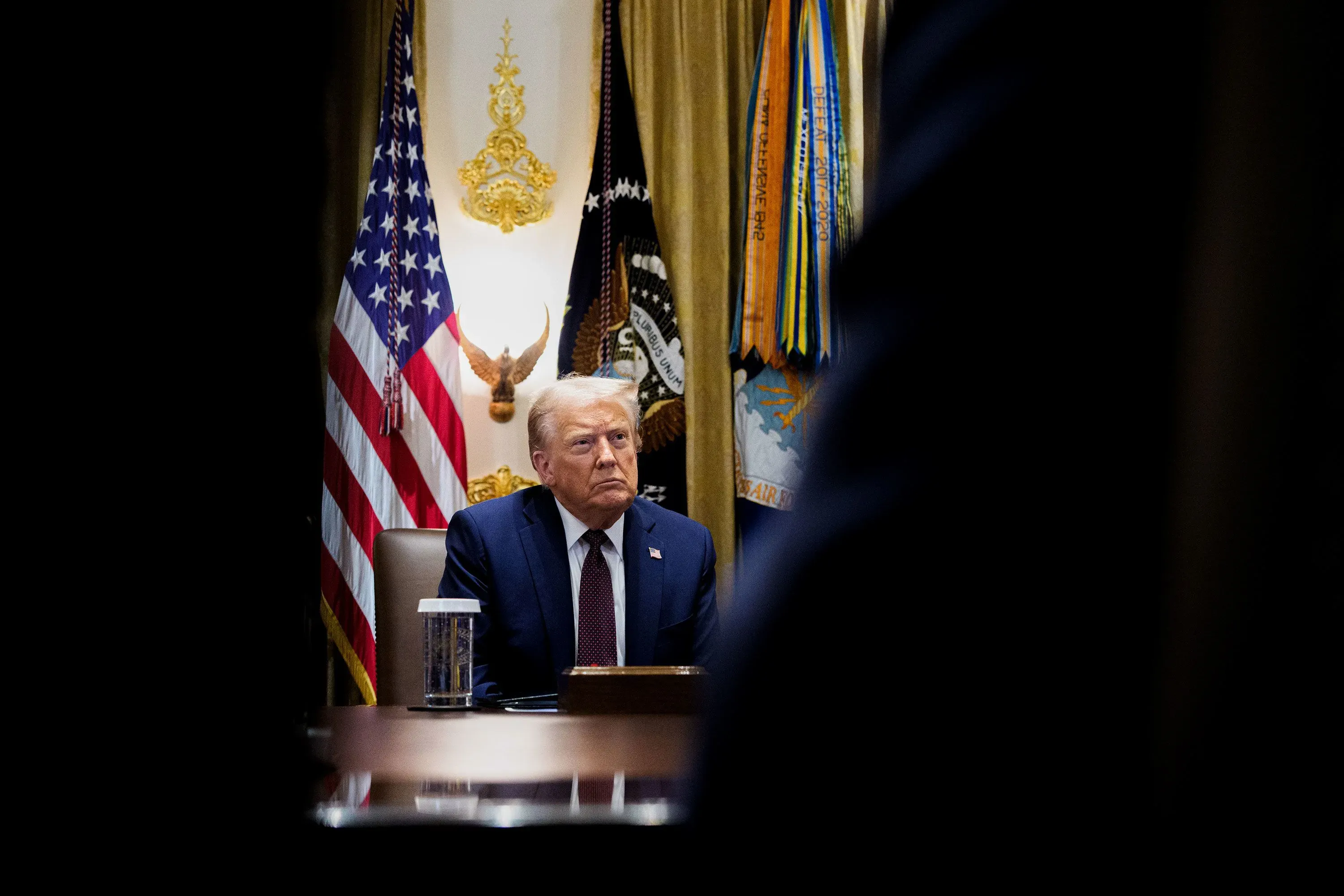 اجتماع في البيت الأبيض يظهر توترًا في فريق ترامب، حيث يجلس الرئيس دونالد ترامب بوجه جدي وسط الأعلام الأمريكية.