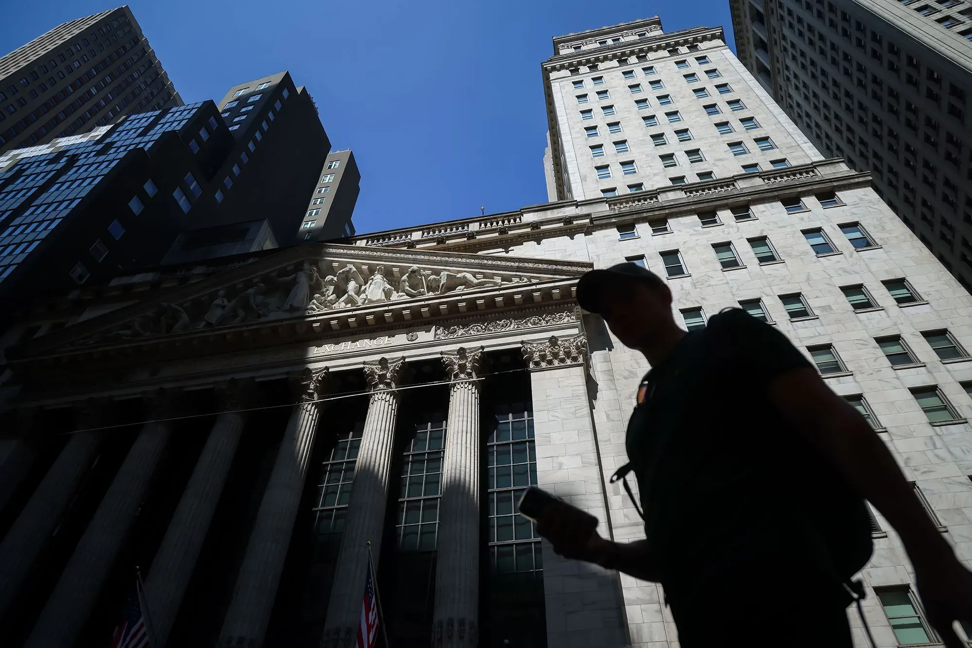 ظل شخص يسير أمام مبنى ضخم في وول ستريت، مع سماء زرقاء وصافية في الخلفية، مما يعكس نشاط السوق المالي.