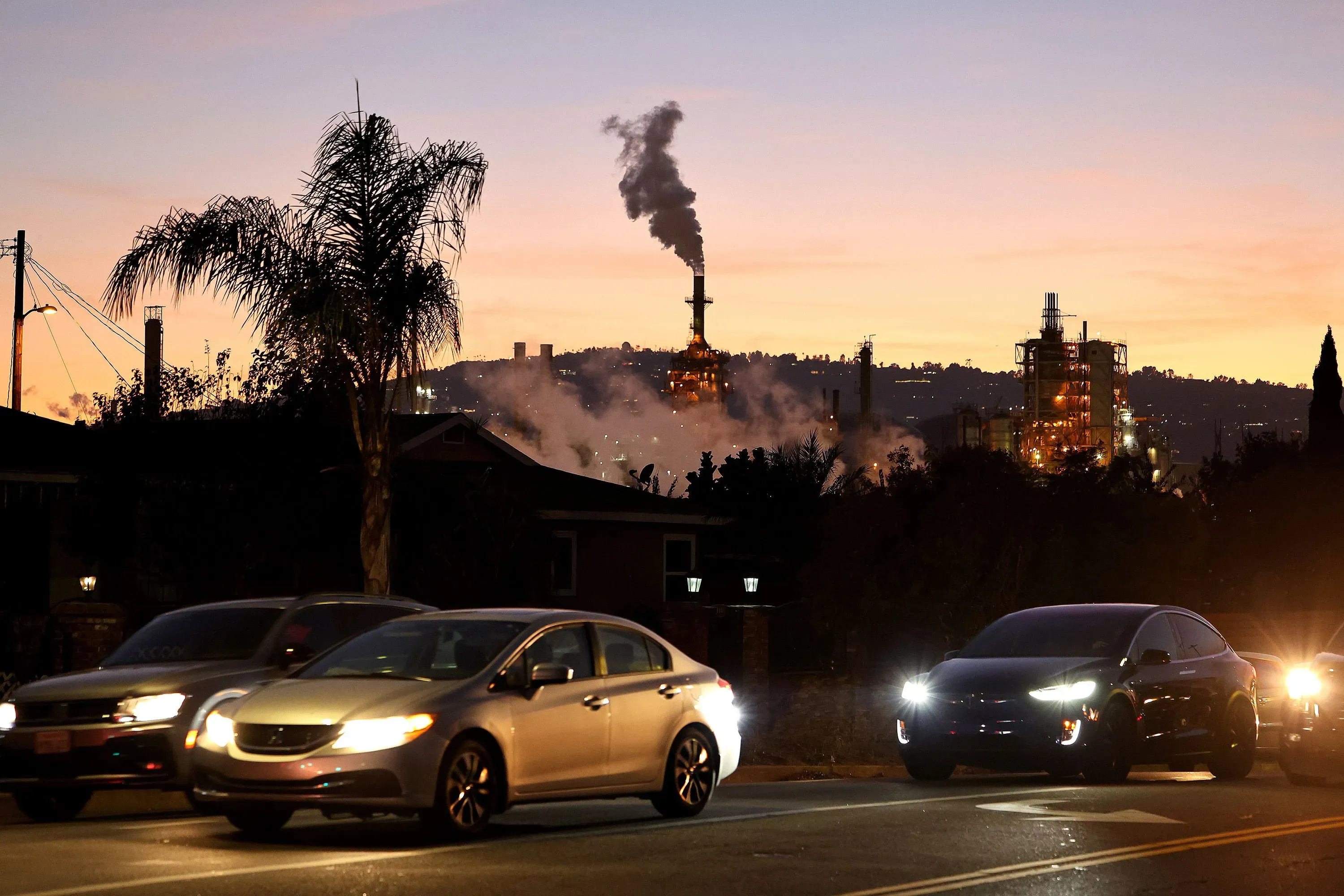 تظهر الصورة مصفاة تكرير في كاليفورنيا مع دخان يتصاعد في السماء، بينما تسير السيارات على الطريق المجاور، مما يعكس التحديات المتعلقة بإمدادات البنزين.