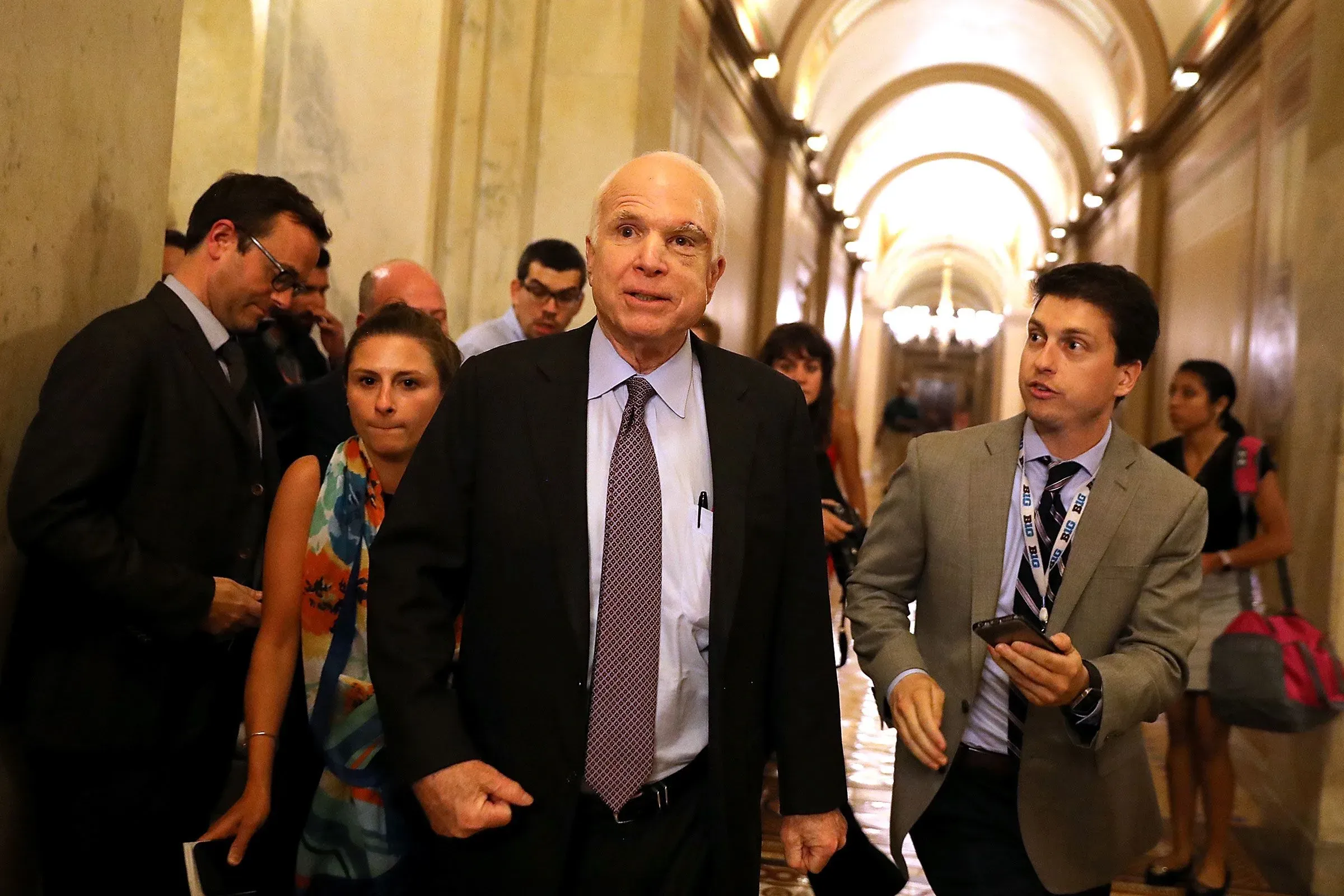 مجموعة من الأشخاص يتحدثون في ممر حكومي، مع ظهور شخصية بارزة في السياسة، مما يعكس التوترات حول قضايا الرعاية الصحية.