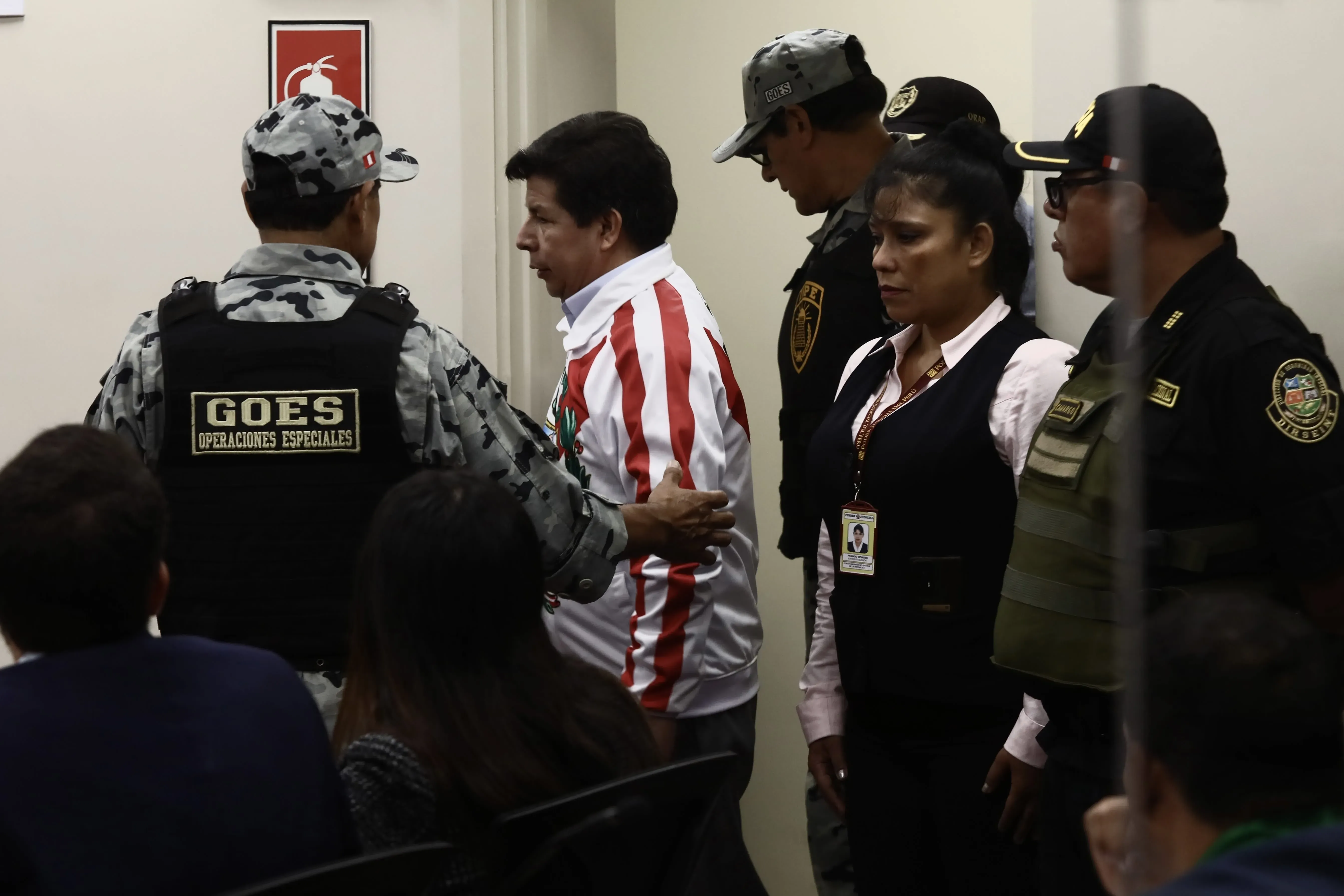 رجل يرتدي زي سجن أبيض مع خطوط حمراء، محاط بضباط أمن أثناء نقله في محكمة بيرو، مما يعكس التحديات القانونية التي تواجه القادة في أمريكا اللاتينية.