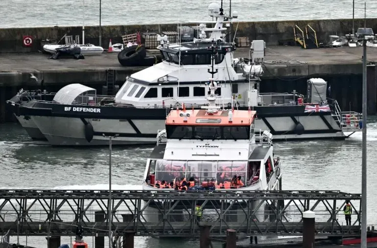 سفينة تحمل مهاجرين تصل إلى ميناء في المملكة المتحدة، مع وجود قارب آخر في الخلفية، في سياق مناقشة حول قوانين الهجرة.