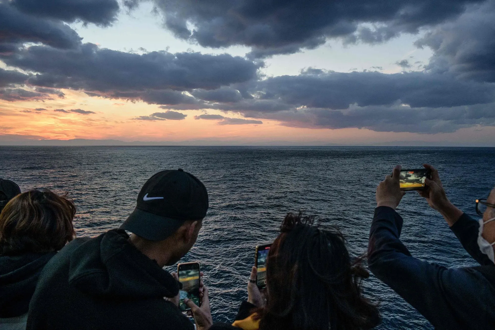 مجموعة من الأشخاص يلتقطون صورًا للغروب باستخدام هواتفهم المحمولة، مع منظر للبحر والغيوم في الأفق.