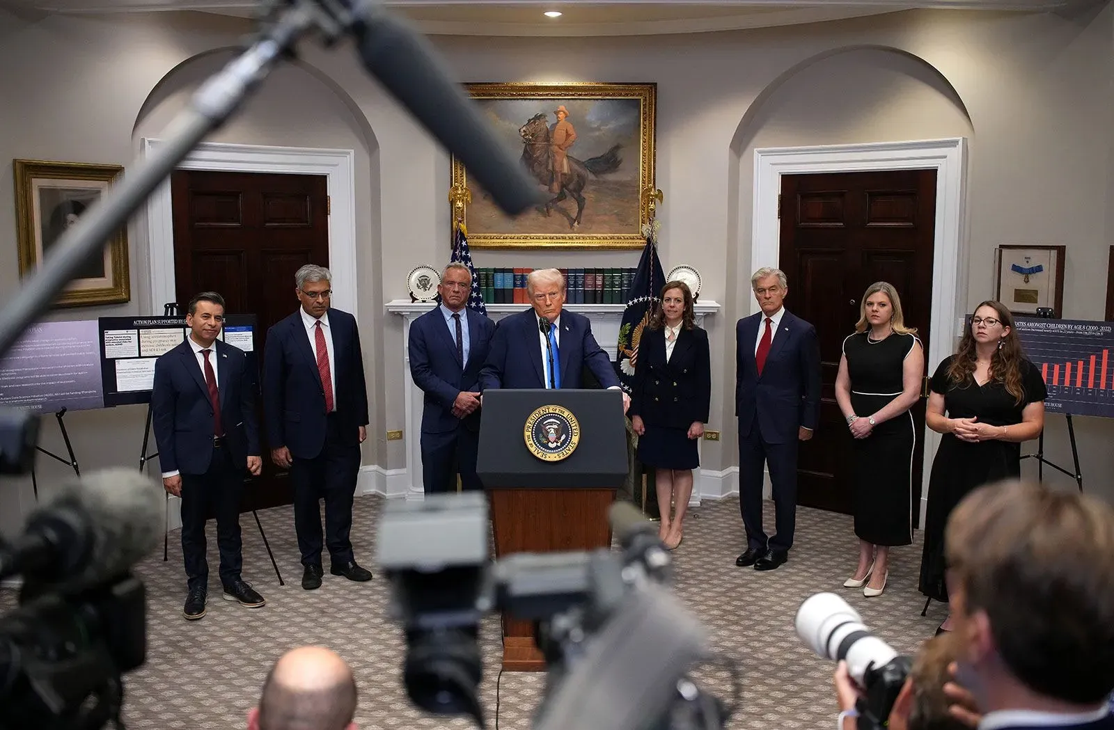 ترامب يتحدث في مؤتمر صحفي بالبيت الأبيض، محاطًا بمساعديه، مع وجود رسومات بيانية خلفهم توضح بيانات مهمة.