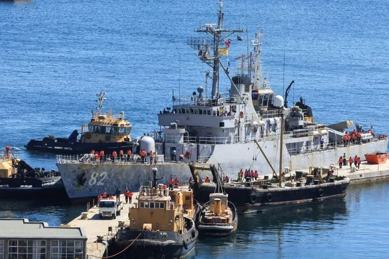 سفينة حربية راسية في ميناء بجنوب أفريقيا خلال مناورات بحرية لدول البريكس، مع وجود قوارب مساعدة وطاقم يعمل على الأرصفة.
