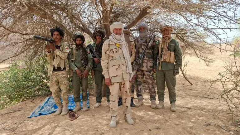 مجموعة مسلحة من جيش تحرير أزواد في شمال مالي، يظهرون بأسلحة تحت ظل شجرة في منطقة صحراوية، تعبيراً عن التحالف العسكري غير المسبوق في الصراع المالي.