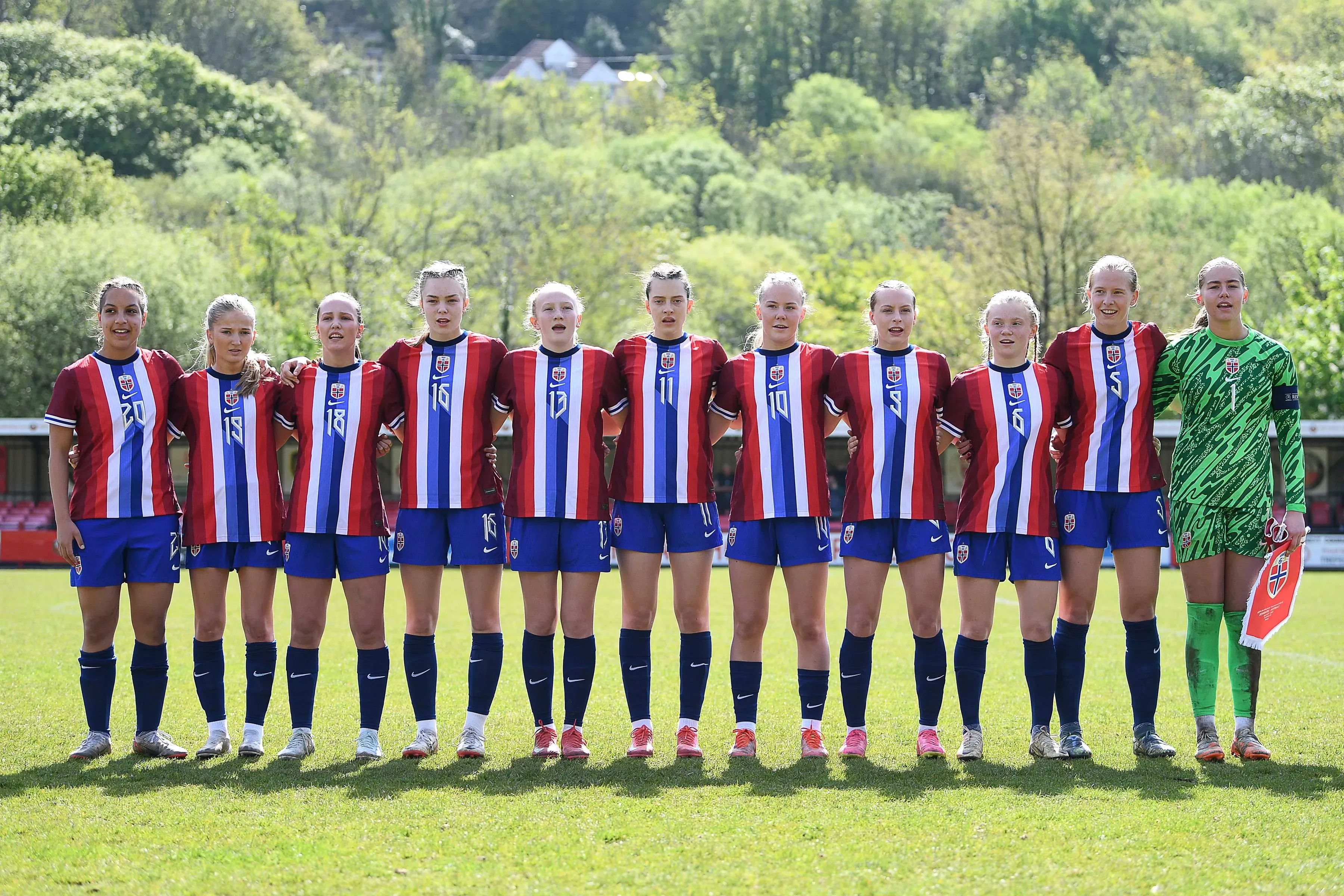 فريق كرة القدم النسائي النرويجي يقف معًا على أرض الملعب، يرتدون قمصانًا حمراء وزرقاء، مع خلفية طبيعية خضراء.