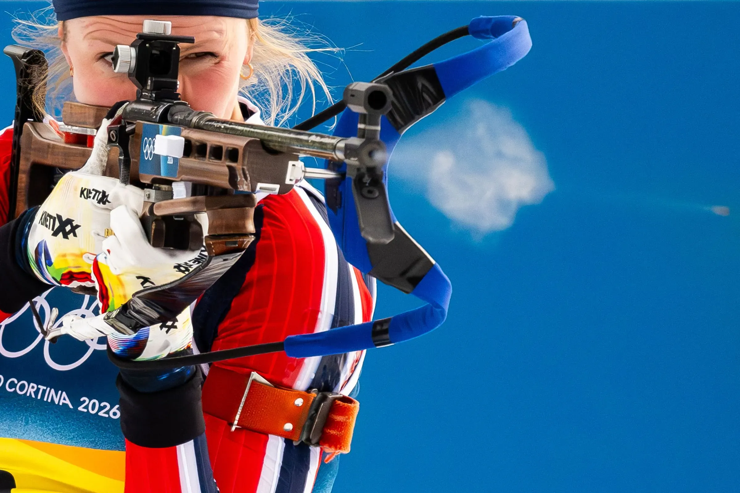 رياضية نرويجية ترتدي زي المنافسة، تستعد لإطلاق النار من بندقية في حدث رياضي أولمبي، مع خلفية زرقاء.