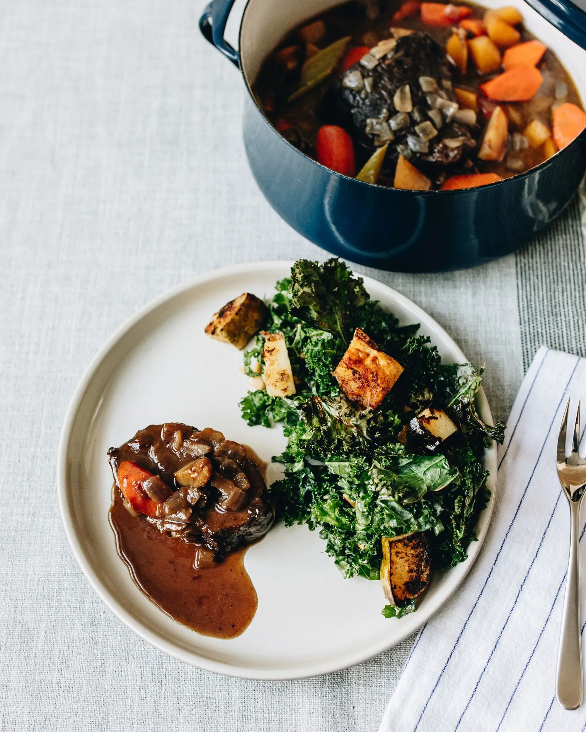 طبق من اللحم المطبوخ مع صلصة غنية، يقدم بجانب سلطة الكالي المحمصة، مع وعاء من الحساء في الخلفية.