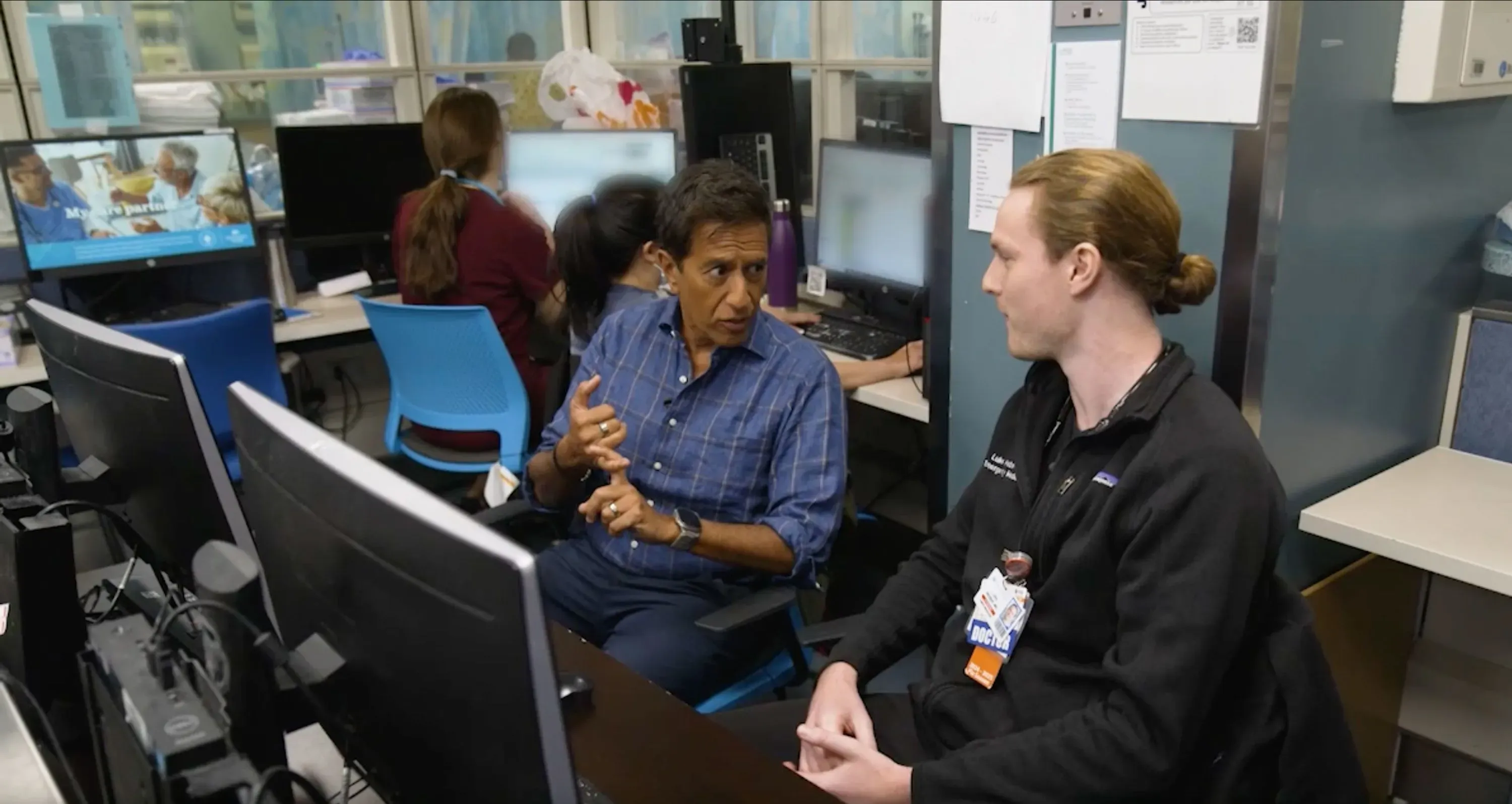 دكتور سانجاي غوبتا يتحدث مع زميله في مركز طبي، مع شاشات كمبيوتر تعرض معلومات طبية خلفهم، مما يعكس أهمية البحث في تخفيف الألم.