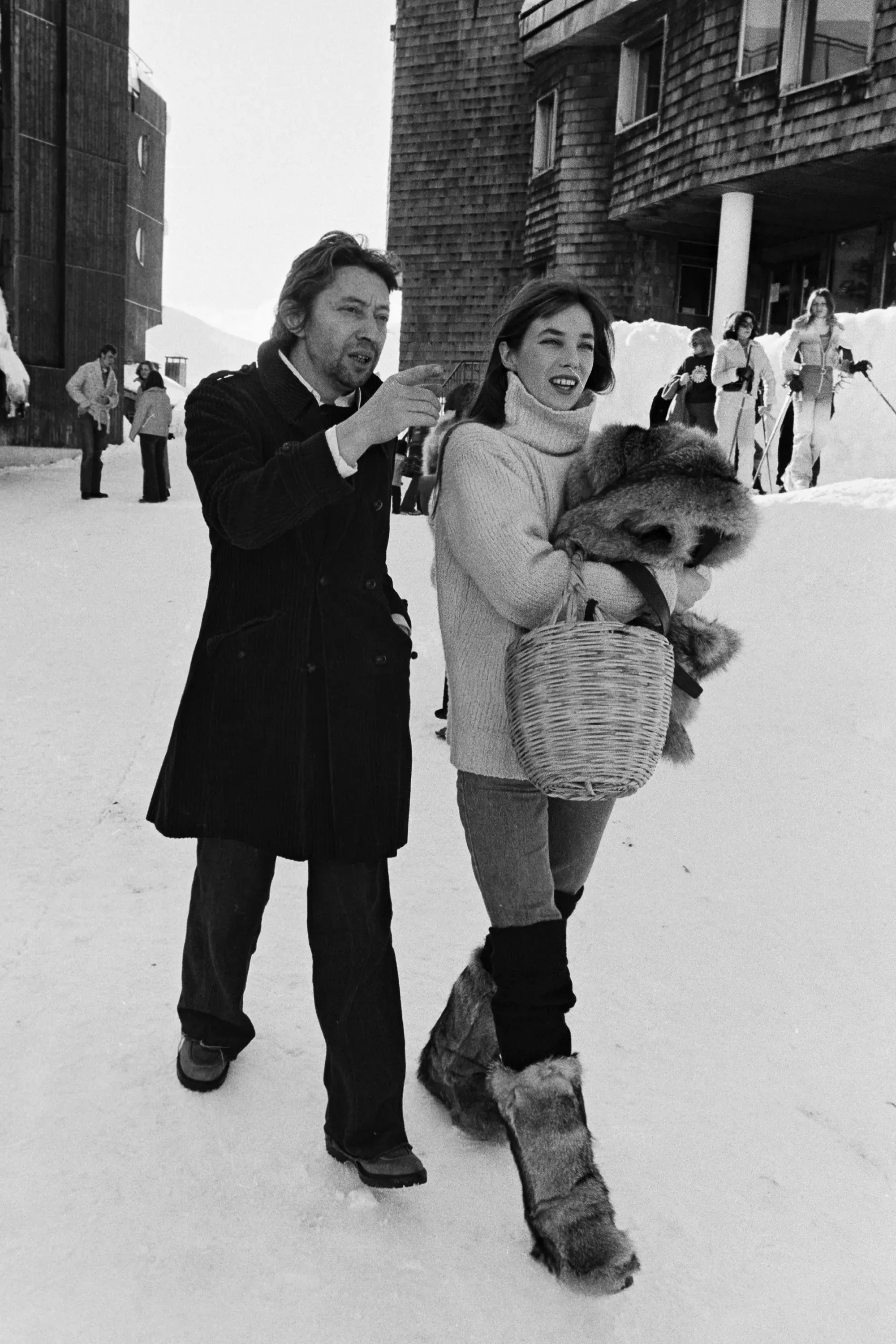 زوجان يتجولان في منتجع تزلج، يرتديان ملابس شتوية أنيقة، مع خلفية من الثلوج والمباني الخشبية، تعكس ثقافة التزلج الفاخرة.