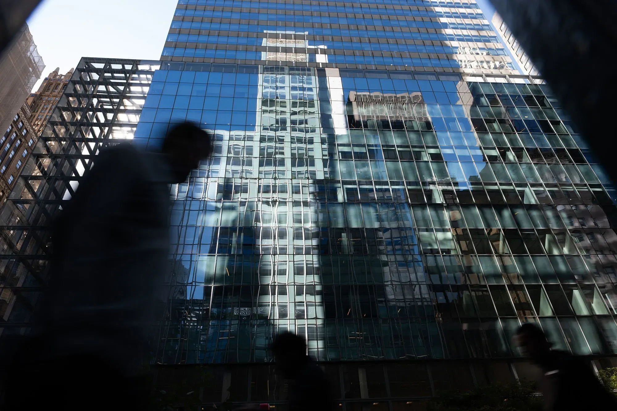ظلال أشخاص يمرون أمام ناطحة سحاب زجاجية تعكس مشهد المدينة، تعبيراً عن تأثير الذكاء الاصطناعي على الأسواق المالية.