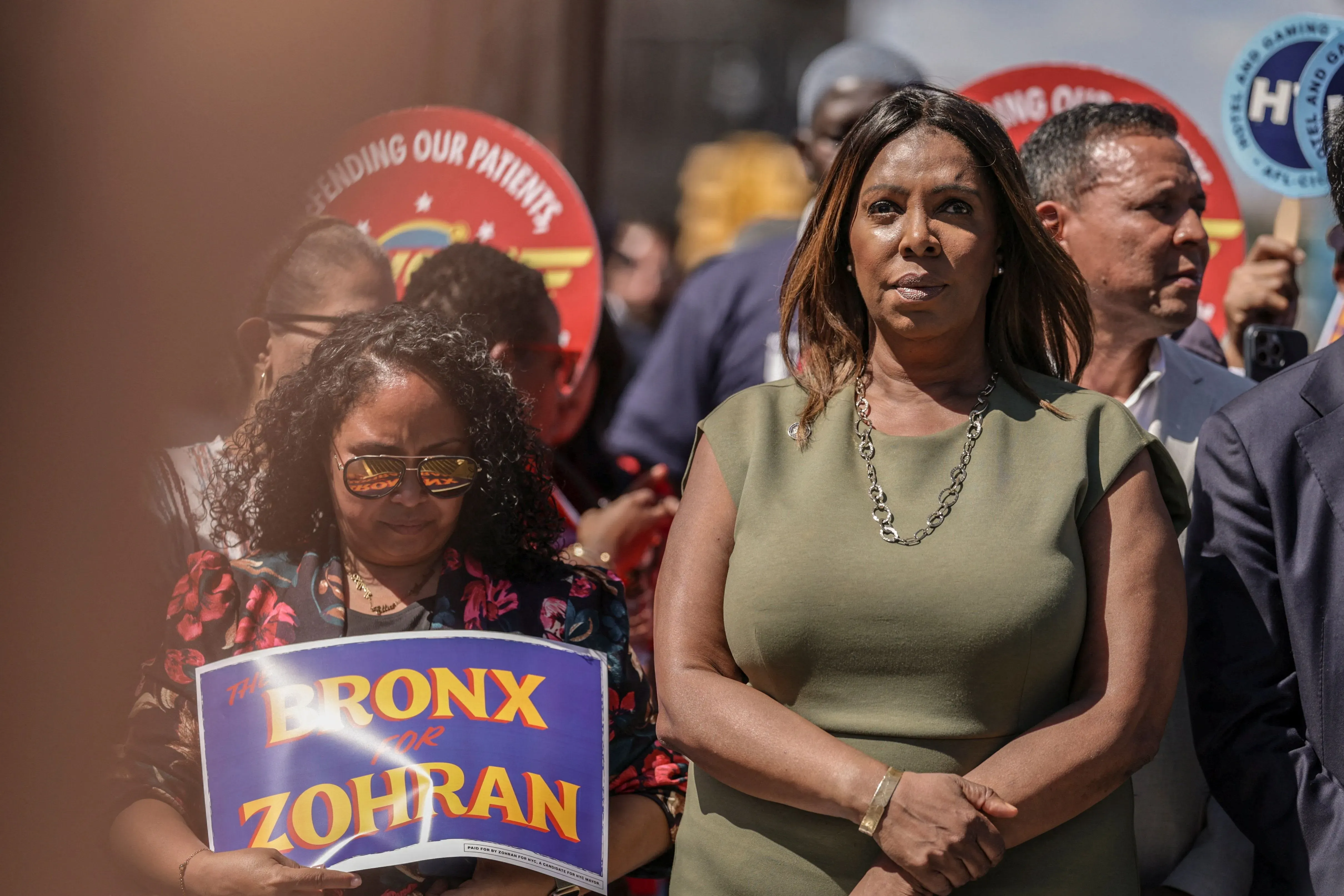امرأة ترتدي فستاناً أخضر وتظهر بوضوح في مقدمة الصورة، بينما تحمل امرأة أخرى لافتة مكتوب عليها "THE BRONX FOR ZOHRAN"، مع مجموعة من الأشخاص في الخلفية.