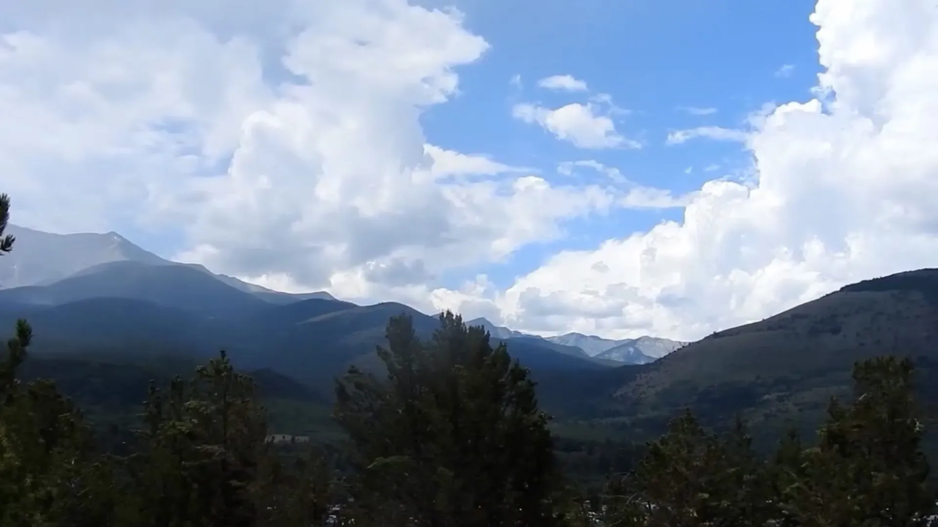 منظر جبلي في مونتانا يظهر الغابات الكثيفة والجبال تحت سماء زرقاء مع غيوم، يسلط الضوء على التحديات التي تواجه السلطات في البحث عن المشتبه به.