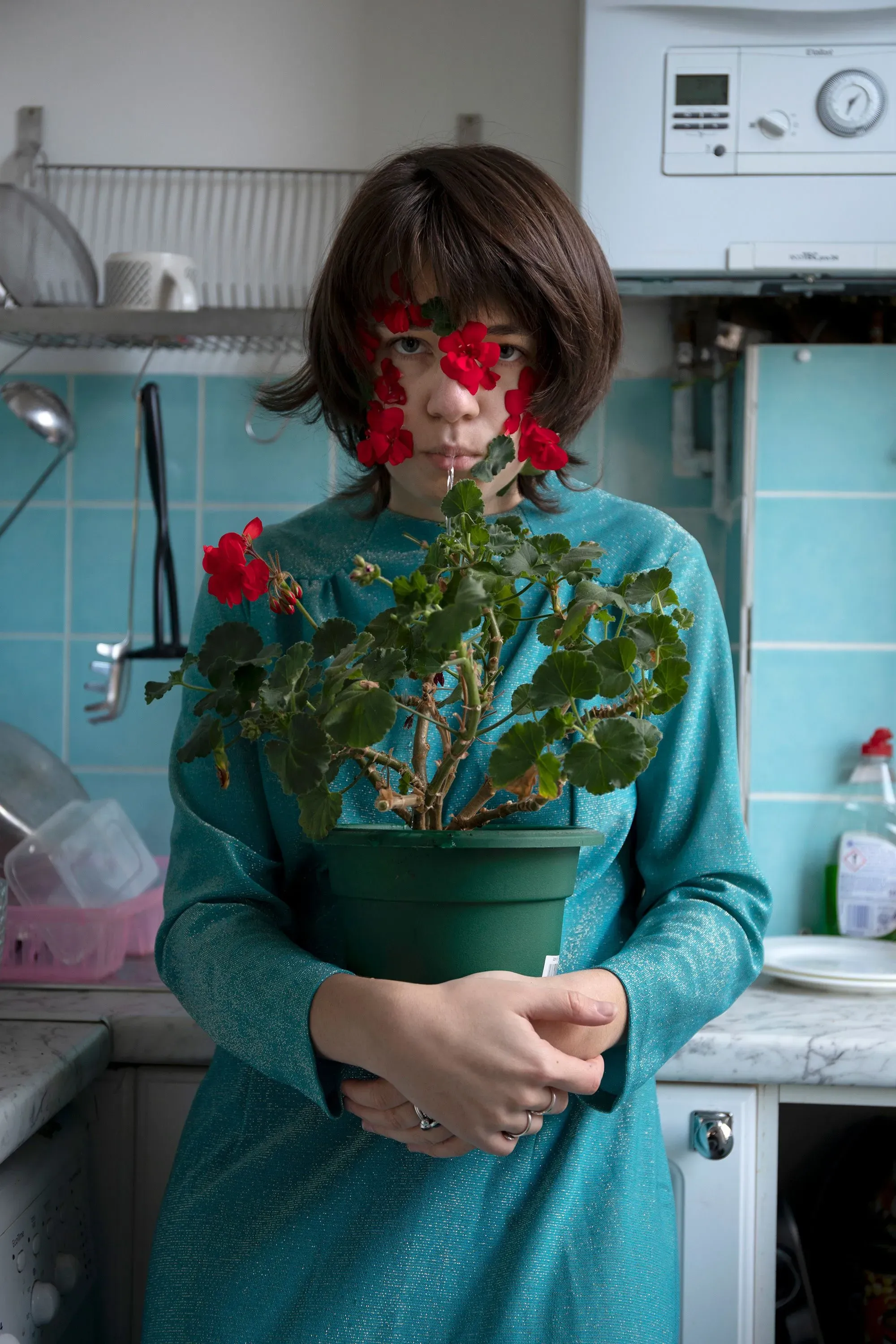 امرأة ترتدي فستاناً أزرق، تحمل نباتاً مزهراً في مطبخ منزلي. تعكس الصورة التحول في تصوير الأزياء نحو البيئات الحميمة.