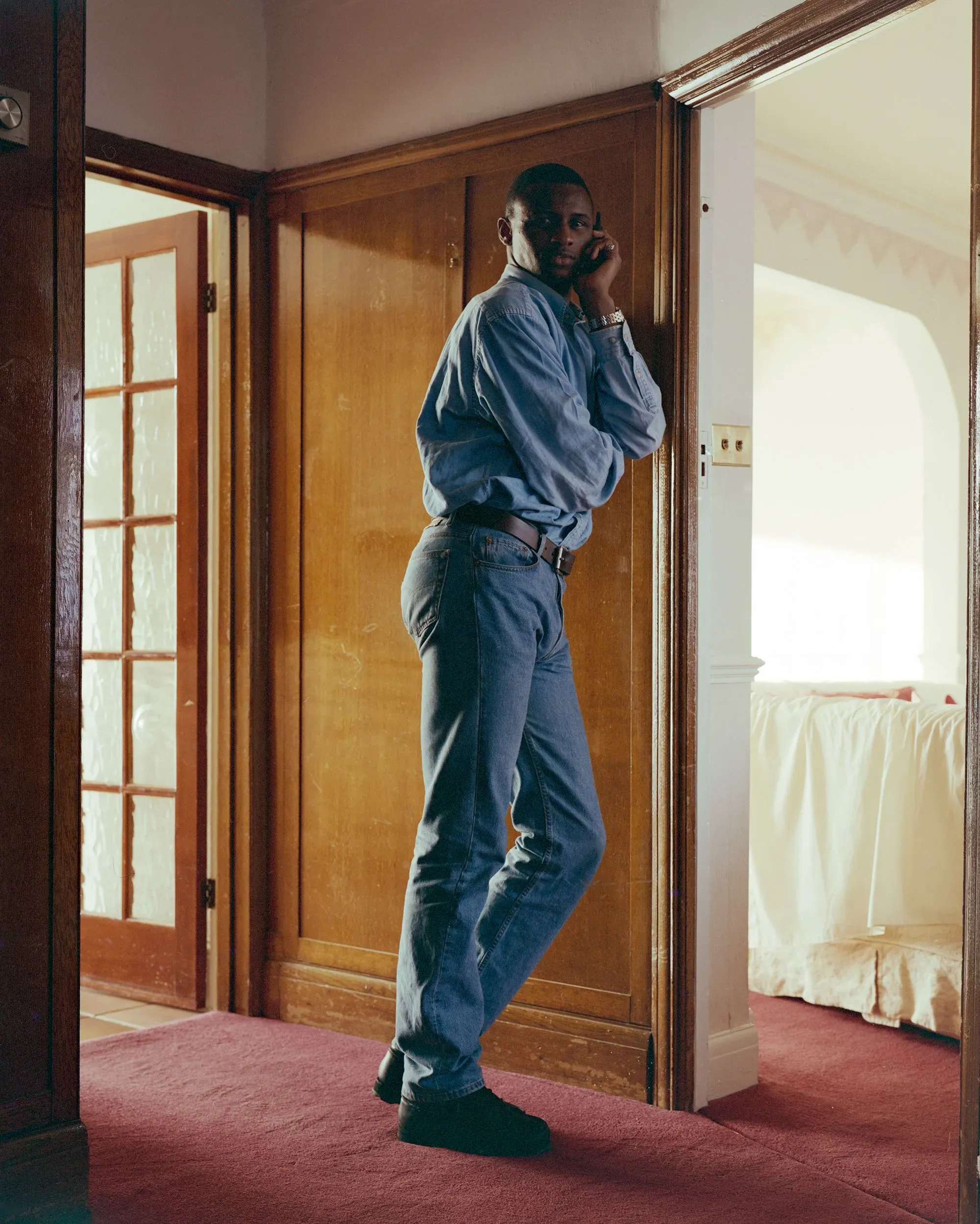 رجل يرتدي قميصًا أزرق وسروال جينز يقف في مدخل منزل، يتحدث في الهاتف، محاط بأجواء منزلية دافئة.
