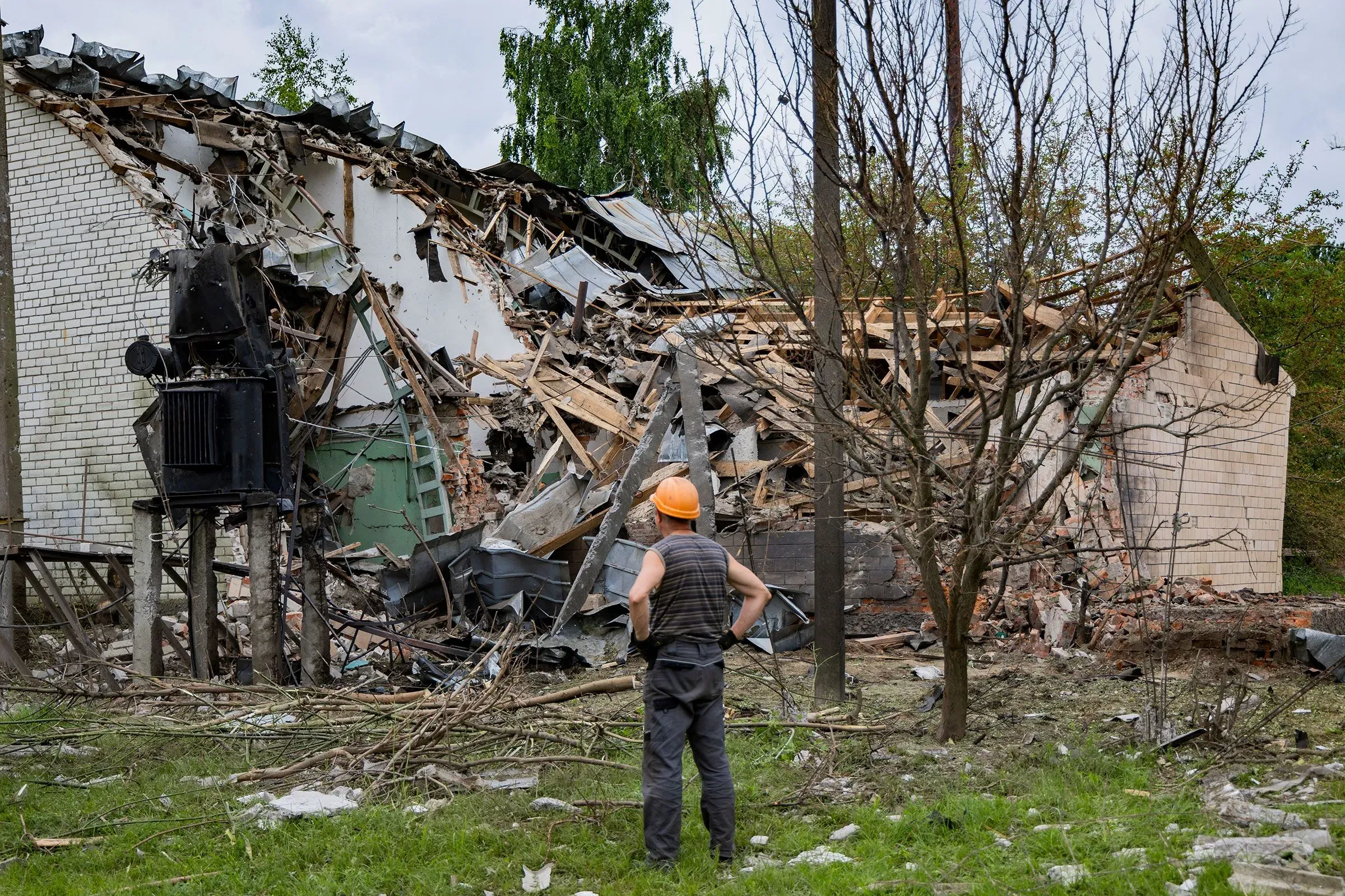 رجل يرتدي خوذة يتأمل حطام مبنى مدمر في أوكرانيا، مما يعكس آثار الهجمات الروسية على البنية التحتية المدنية.