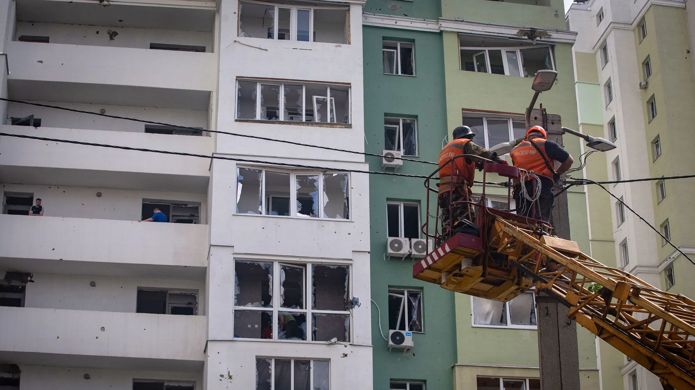 عمال يقومون بإصلاح أعمدة الكهرباء بالقرب من مبنى سكني متضرر في أوكرانيا، مما يعكس جهود إعادة بناء البنية التحتية للطاقة.