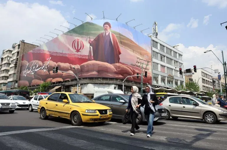 لوحة إعلانات ضخمة في طهران تظهر شخصية بارزة ترتدي عباءة، بينما يعبر الناس الشارع وسط حركة المرور. تعكس الصورة التوترات الاقتصادية والسياسية في المنطقة.