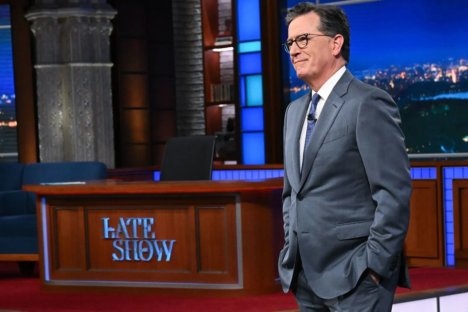 ستيفن كولبرت يقف على المسرح في برنامجه "The Late Show"، مع خلفية مضاءة وأثاث استوديو مميز، وسط أجواء من التوتر بعد قرارات الشبكات التلفزيونية.
