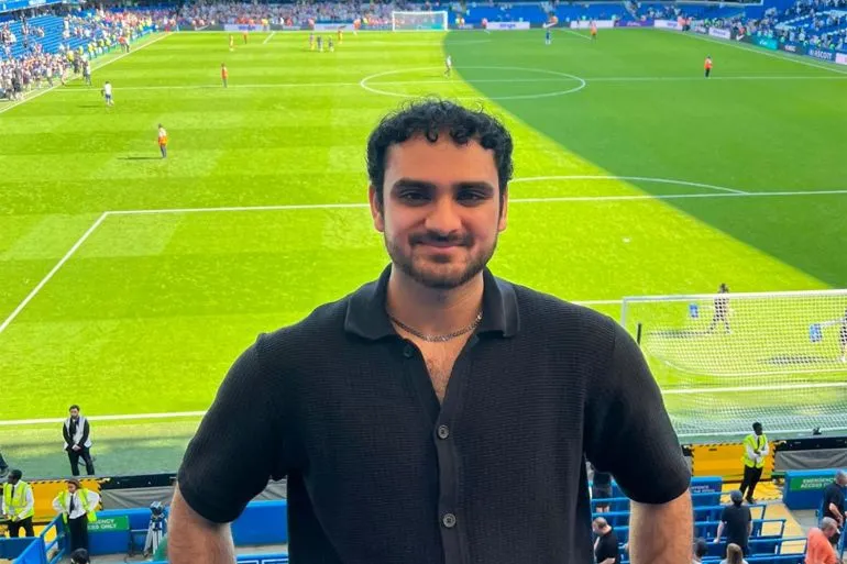 شاب مسلم مبتسم يقف في مدرجات ملعب كرة قدم خلال مباراة كأس العالم، مع خلفية الملعب الأخضر والجماهير.