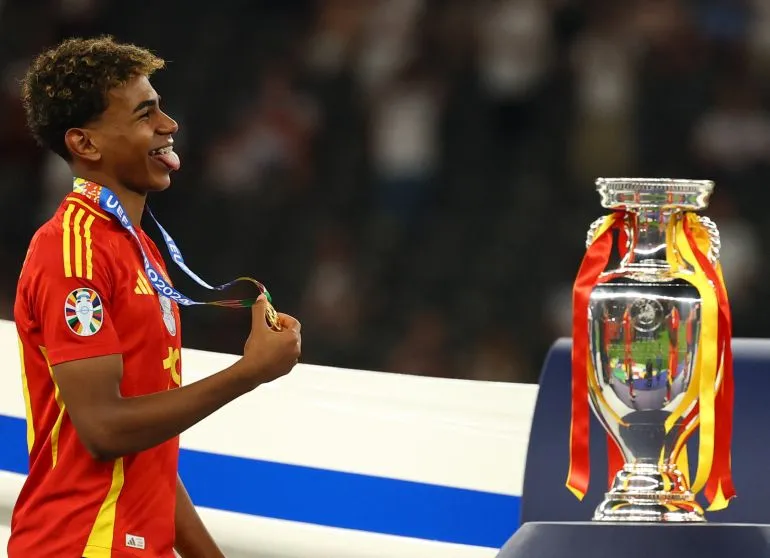 لامين يامال يحتفل بفوزه مع المنتخب الإسباني بكأس أوروبا 2024 مرتدياً الميدالية الذهبية بجانب الكأس الرسمية.