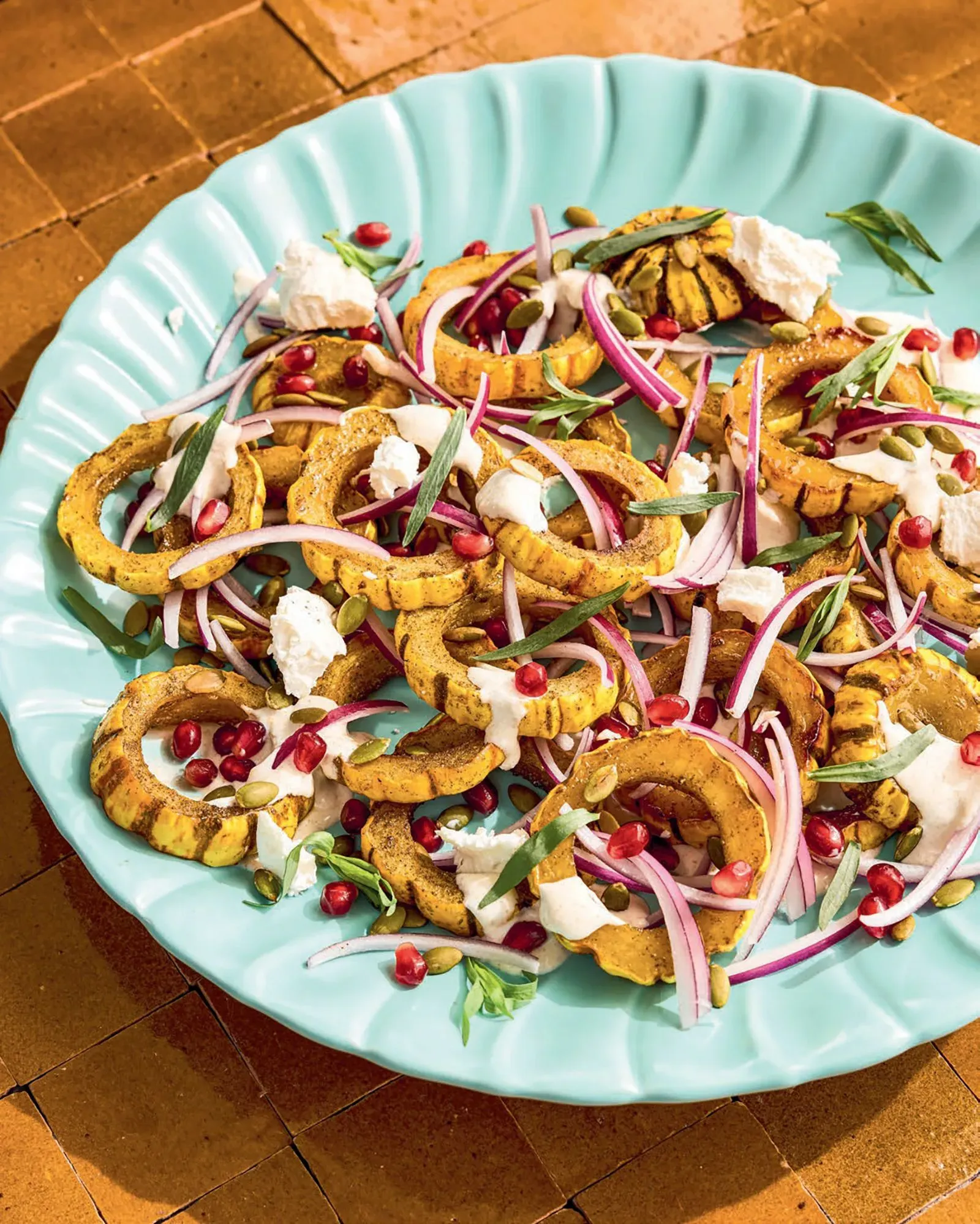 سلطة ملونة تضم شرائح القرع الداليكاتا المحمص، جبن الماعز، بذور اليقطين، والبصل الأحمر، موضوعة على طبق زينة أزرق.