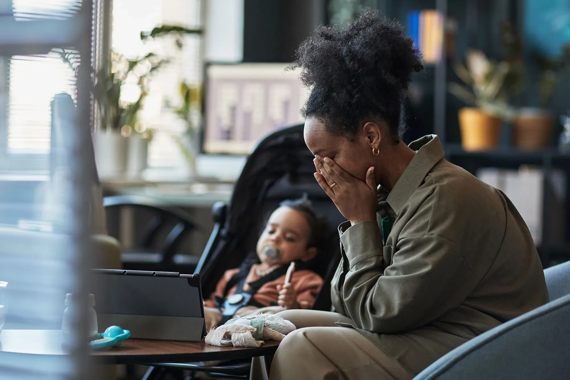 امرأة تعبر عن مشاعر القلق والإرهاق أثناء جلوسها في مكان عام، بينما يجلس طفلها في عربة قريبة.
