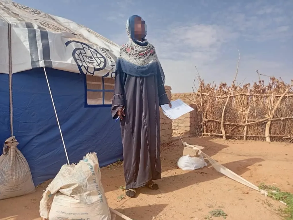 امرأة سودانية تقف أمام خيمة إغاثة في منطقة صحراوية، تعكس تأثير النزاع المسلح في مدينة الأبيض على المدنيين واللاجئين.