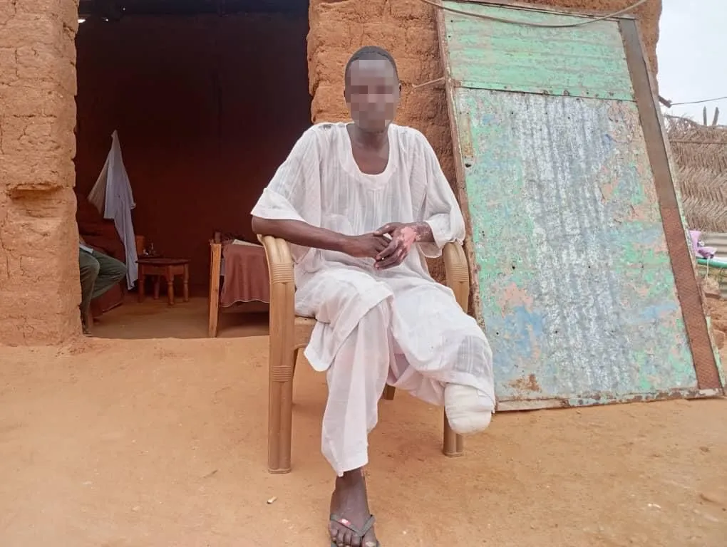 شاب سوداني جالس أمام منزل طيني، يرتدي ملابس بيضاء، ويعاني من بتر في ساقه اليسرى جراء هجوم طائرة مسيّرة في مدينة الأبيض.
