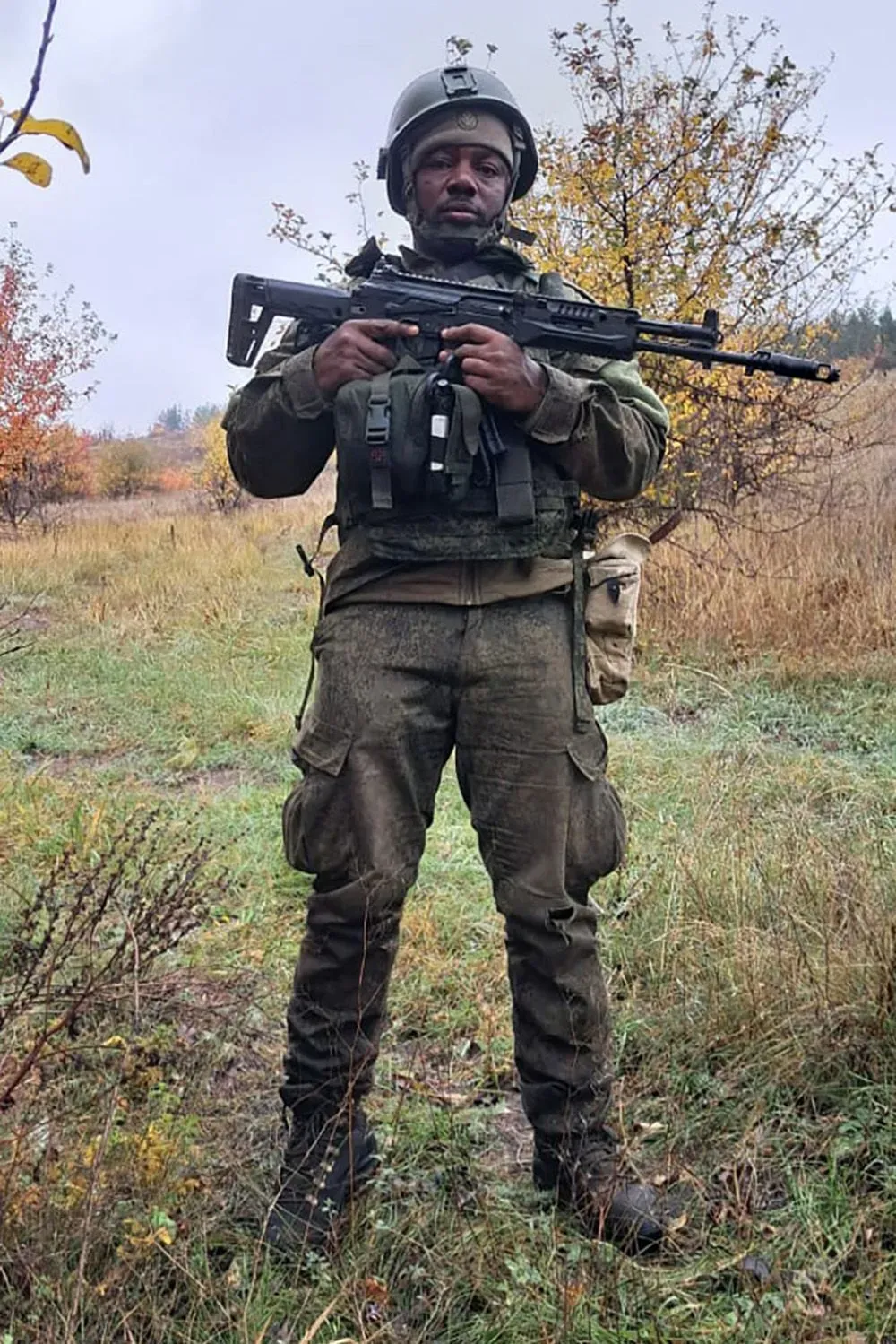 فرانسيس ندونغو، مقاتل أفريقي يرتدي زيًا عسكريًا ويحمل سلاحًا، يقف في منطقة مفتوحة، مع خلفية طبيعية.