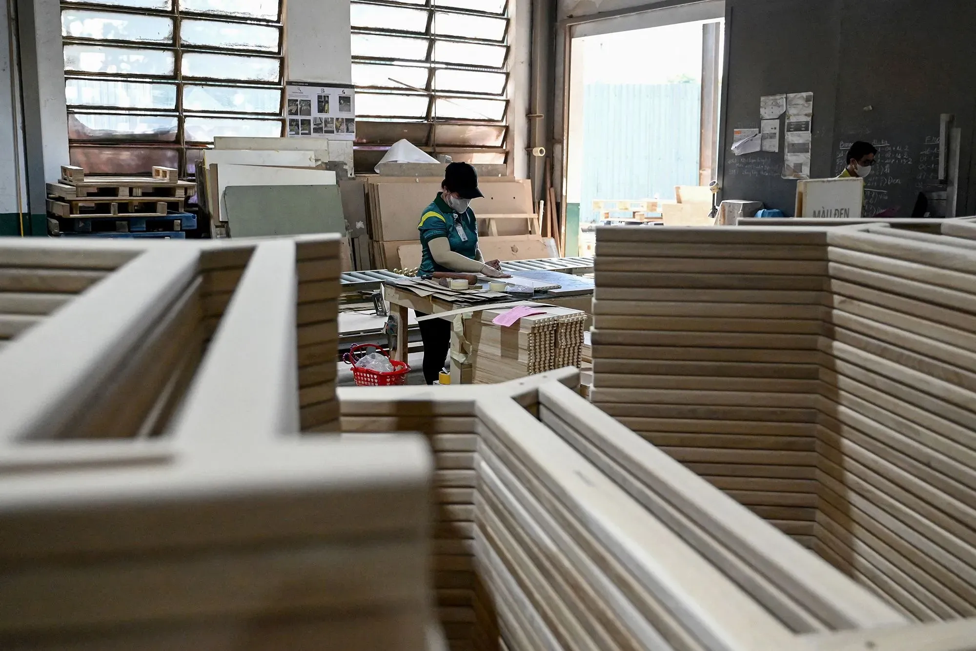 مشهد لمصنع حيث يعمل شخص على تجهيز الأثاث الخشبي، مع وجود قطع خشبية مرتبة في الخلفية، مما يعكس تأثير التعريفات الجمركية على الأسعار.