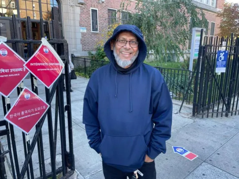 ممداني مبتسم يرتدي سترة زرقاء، يقف أمام لافتات انتخابية متنوعة في نيويورك، معبرًا عن تفاؤله بفوزه التاريخي كعمدة.