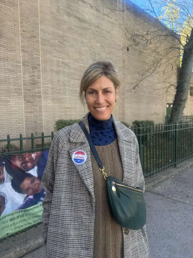 امرأة مبتسمة ترتدي سترة بنمط مربع وشارة "لقد صوتت"، تقف أمام جدار بسيط، مما يعكس أجواء الانتخابات في نيويورك.