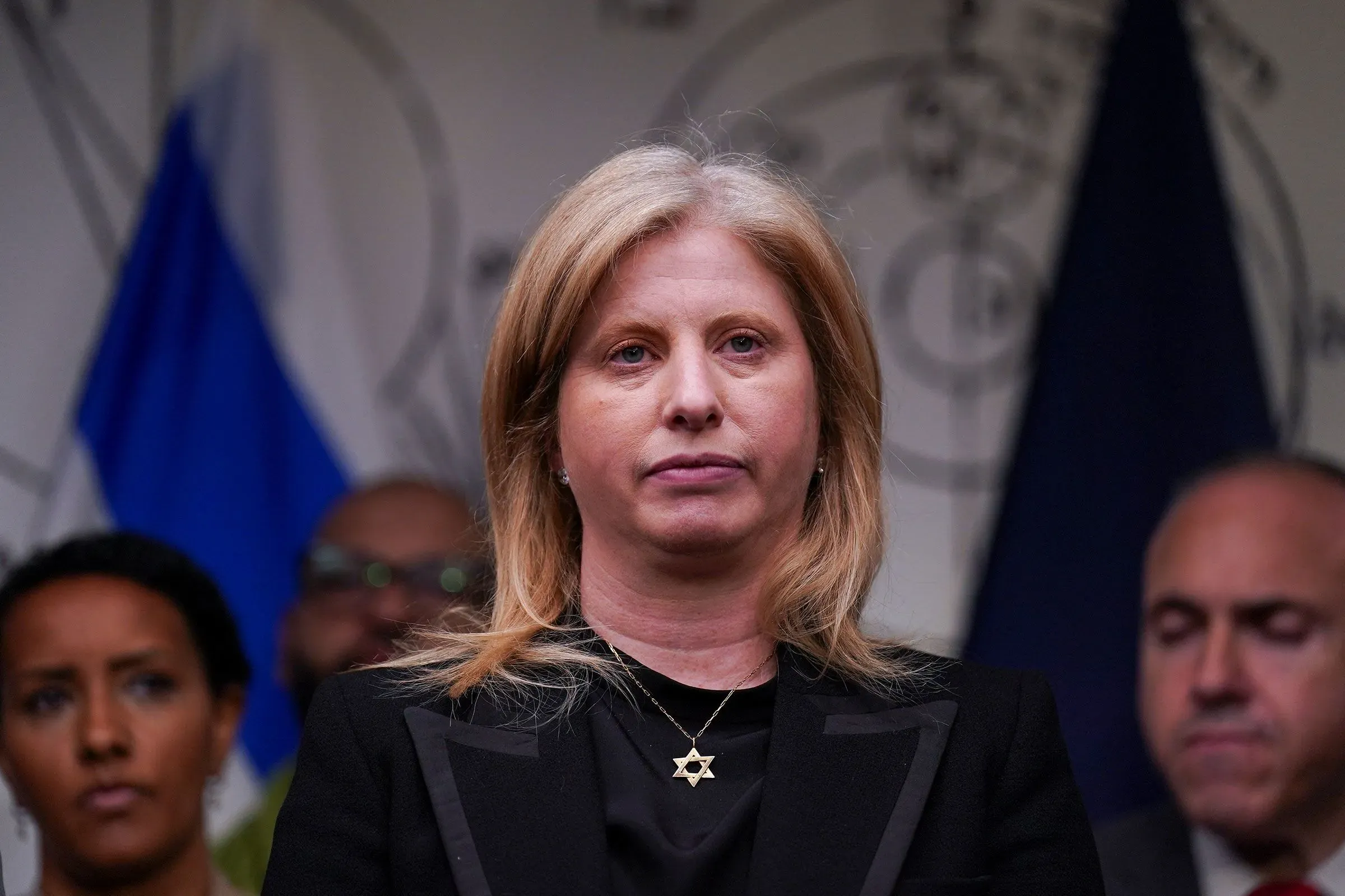 امرأة ترتدي سترة سوداء، تحمل قلادة، وتظهر تعبيرات جدية، بينما يقف خلفها عدد من الأشخاص في مؤتمر صحفي حول قضايا السلامة العامة في نيويورك.