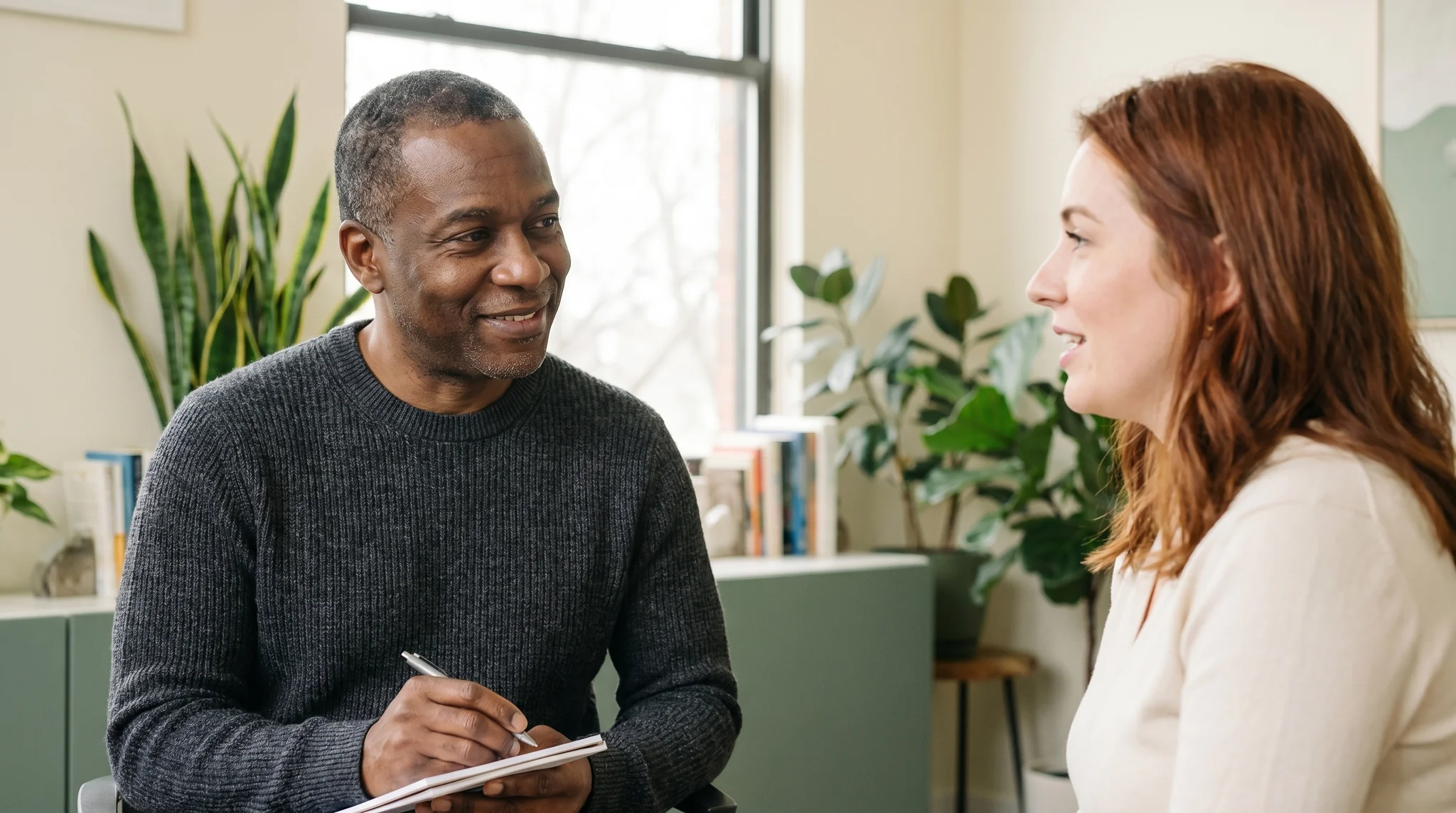 Two people in a supportive one-on-one coaching conversation in a bright office, one taking notes