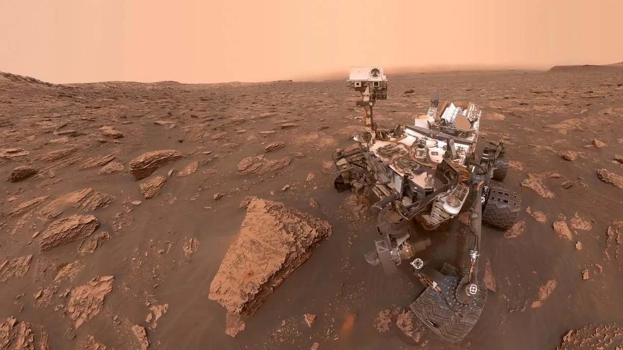 المركبة الجوّالة Curiosity على سطح المريخ وسط تضاريس صخرية حمراء، توضح تقدمها في استكشاف جبل شارب ومسارها الطويل منذ 2012.