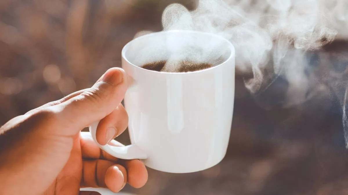 يد تمسك بفنجان أبيض يحتوي على قهوة ساخنة يعلوها بخار، مع التركيز على تأثير رائحة القهوة في تحسين المزاج والاسترخاء بحسب دراسة يابانية حديثة.