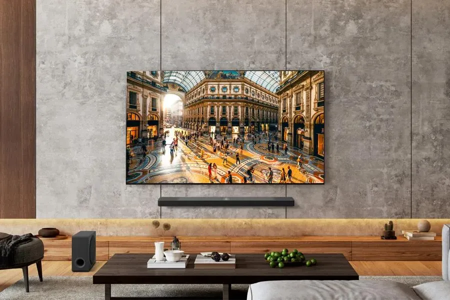 تلفاز ذكي من طراز LG OLED65G3PUA معلق على جدار رمادي في غرفة معيشة حديثة، يعرض صورة مشرقة لسوق داخلي مزدحم.