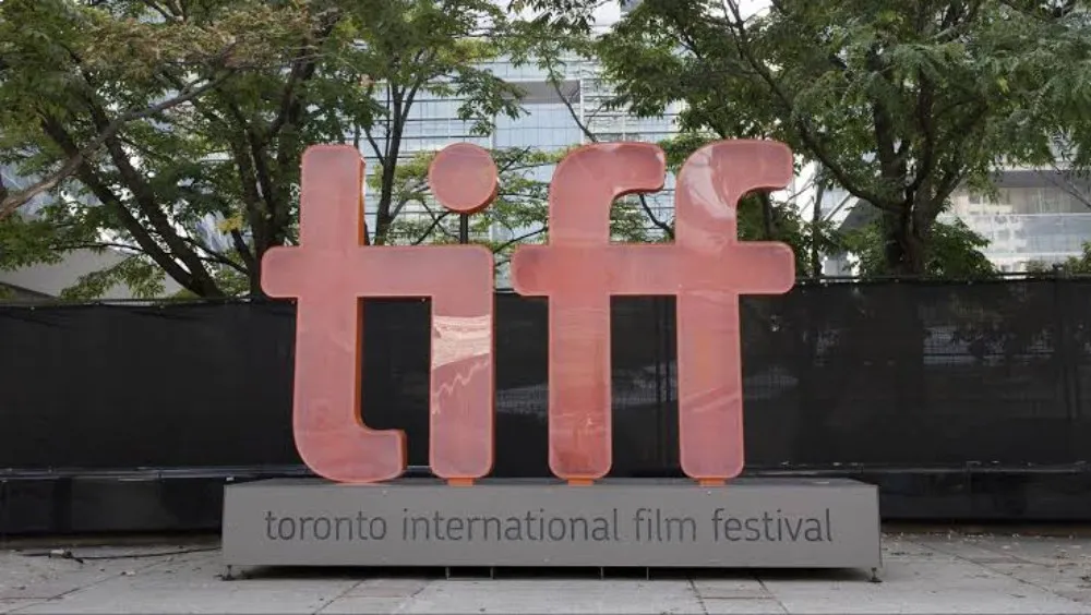 شعار مهرجان تورنتو الدولي للأفلام (TIFF) الكبير باللون الأحمر في موقع خارجي مع أشجار ومباني خلفية، يعكس أهمية المهرجان السينمائي العالمي.